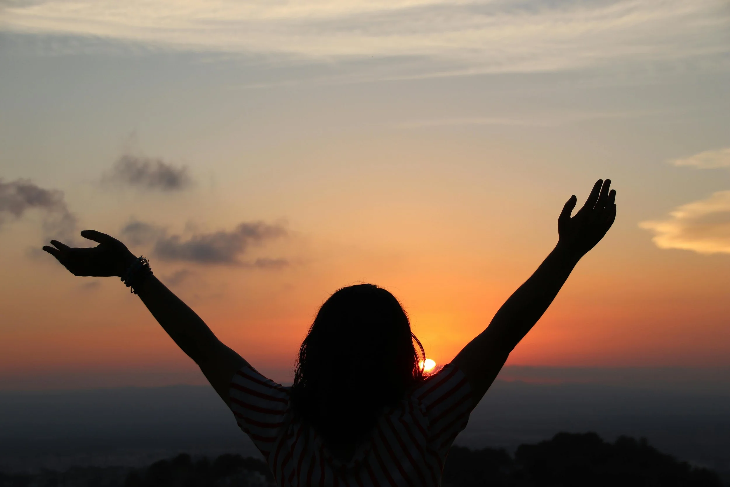 Womans silhouette holding up her hands while overlooking a sunset over the ocean.