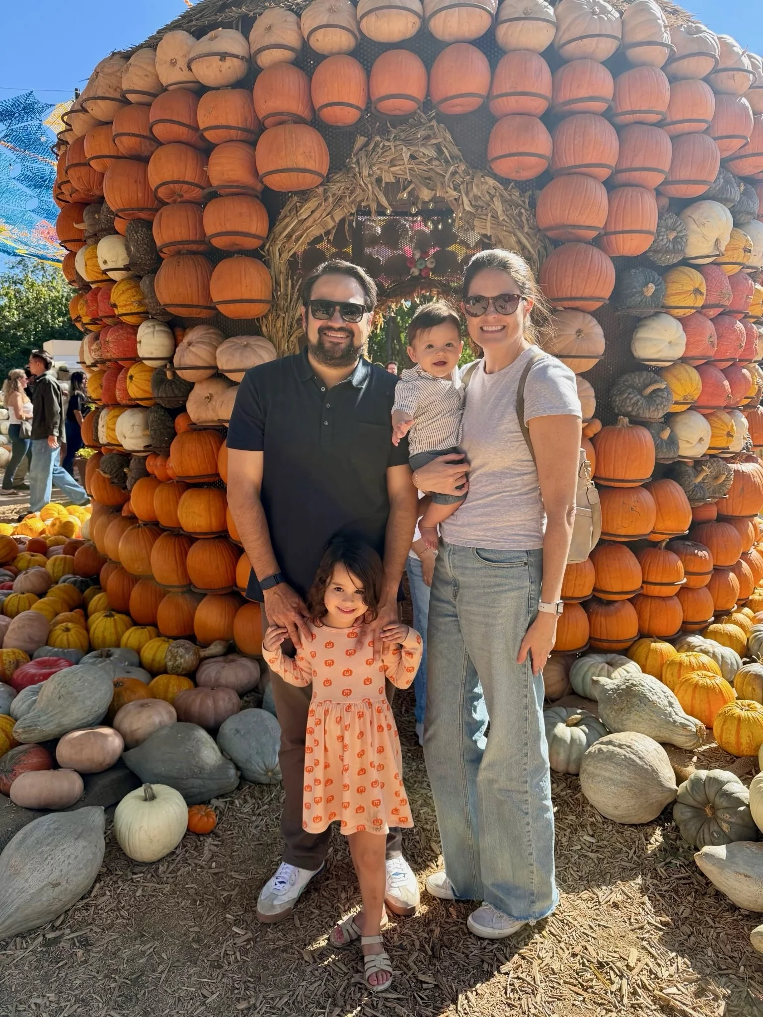 A family of four standing in front of a large pumpkin display at a pumpkin patch. The display is decorated with numerous pumpkins of various colors and sizes, arranged in a large arch with a small pumpkin-shaped house in the center. The family is smiling and dressed casually, with the mother holding a baby and the father holding a young girl.