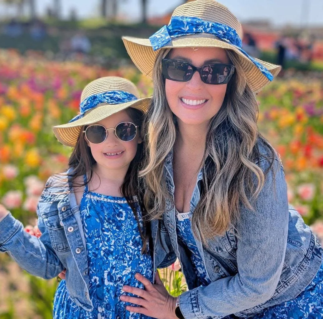 A woman and a girl smiling in a field of colorful flowers, both wearing straw hats with blue ribbons, sunglasses, and blue patterned clothing.