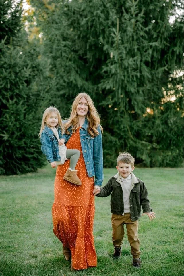 A woman with red hair and two young children, a girl and a boy, walking outdoors on grass with large trees in the background. The woman is holding the girl's hand, and the girl is sitting on the woman's arm. The boy is walking beside them, smiling and holding her hand.