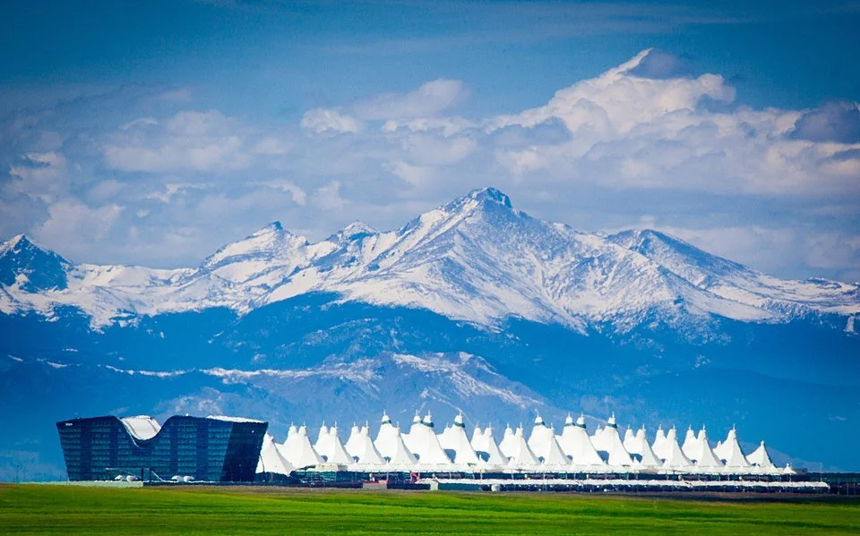 Snow-capped mountains in the background with a modern building and a row of white tents in the foreground on a green landscape.