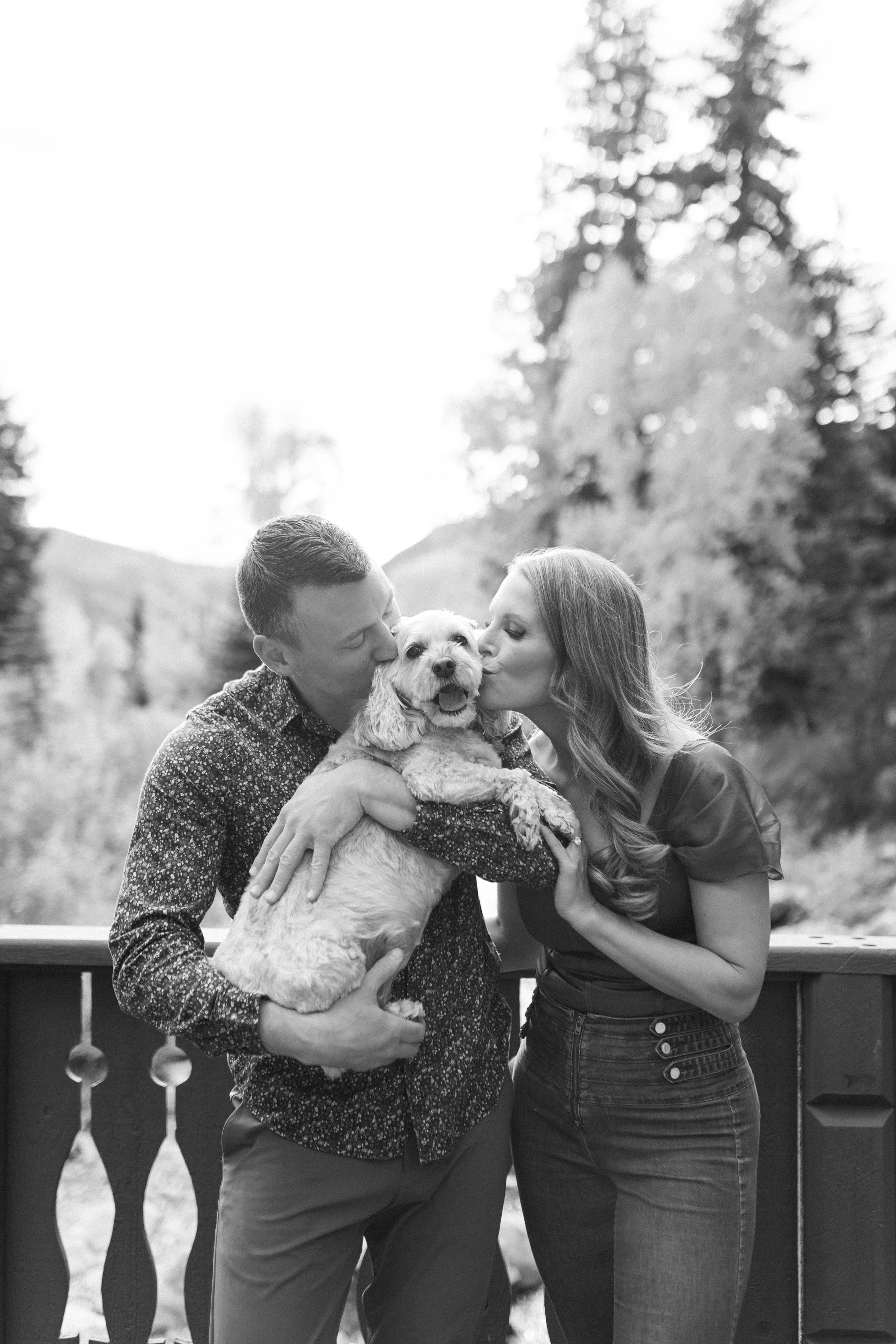 A couple is kissing their dog on the cheeks while holding it, outdoors on a wooden deck with trees and hills in the background.