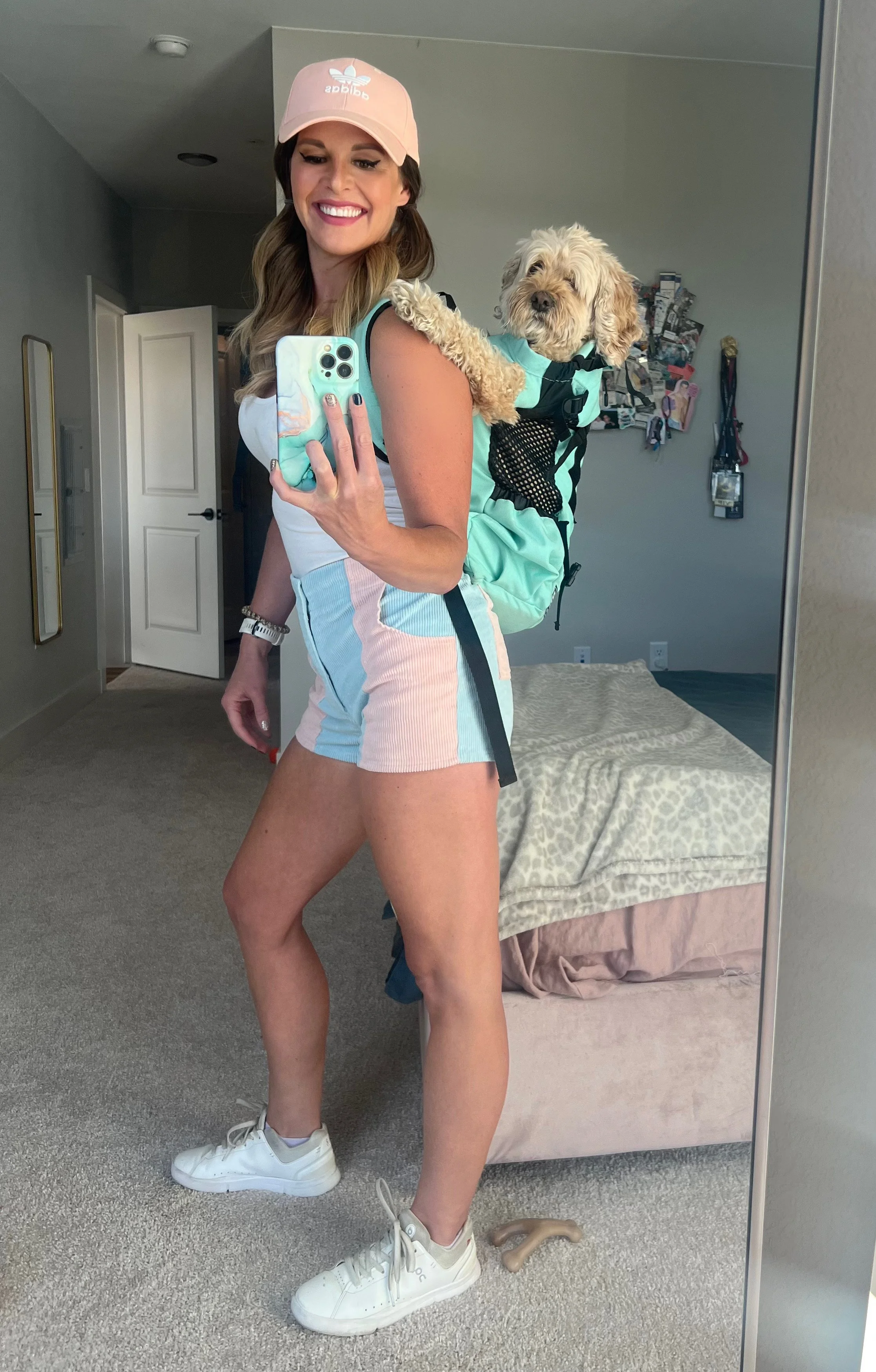 A smiling woman taking a mirror selfie with her smartphone, wearing a pink Adidas cap, a white tank top, colorful shorts, and white sneakers. She has a fluffy dog in a backpack on her back, looking at the camera. The room has a bed with a leopard print blanket and some hanging pictures on the wall.