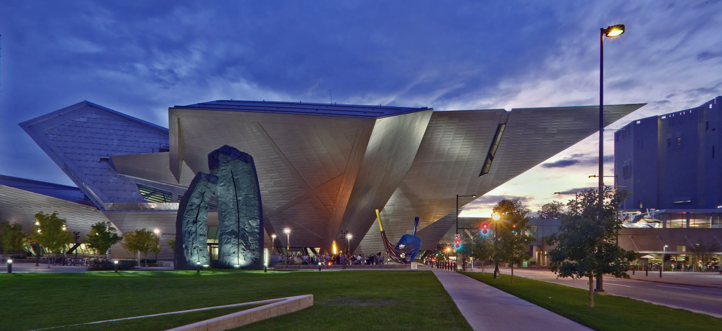 Modern building with angular design illuminated at dusk, surrounded by trees, sculptures, and streetlights.