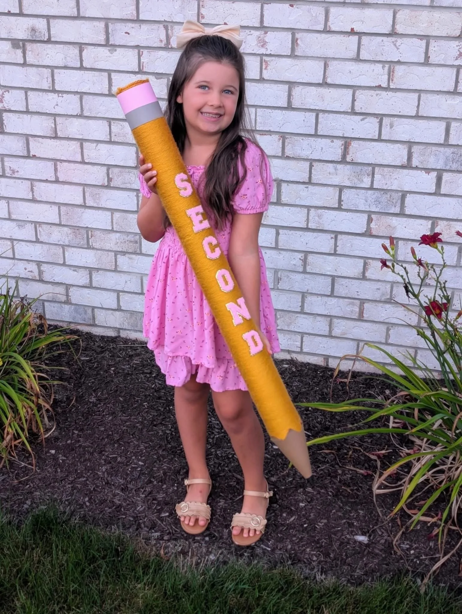 A young girl with long brown hair, wearing a pink dress with ruffled hem, beige heels, and a bow in her hair, standing outdoors in front of a white brick wall. She is smiling and holding a large, yellow, cone-shaped object decorated with pink and white letters that spell "SECOND." The object appears to be a giant pencil or crayon.