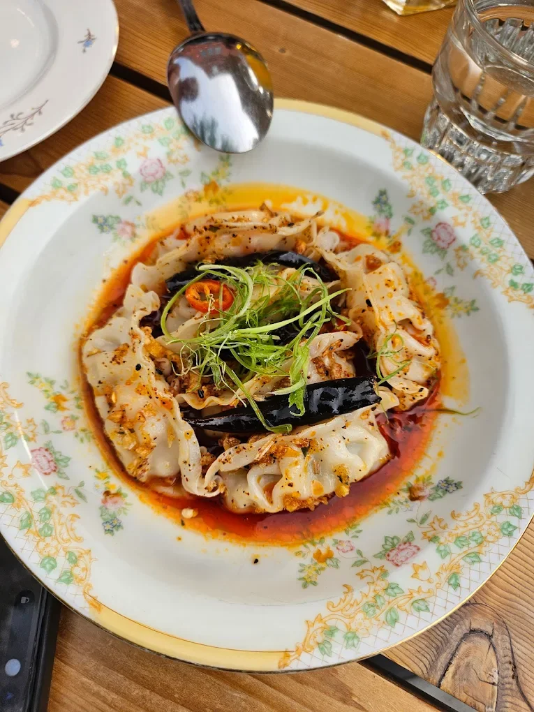 A decorative white porcelain plate with floral patterns, containing Asian dumplings in a spicy red sauce, garnished with green herbs and red chili slices, placed on a wooden table alongside a glass of water and a spoon.