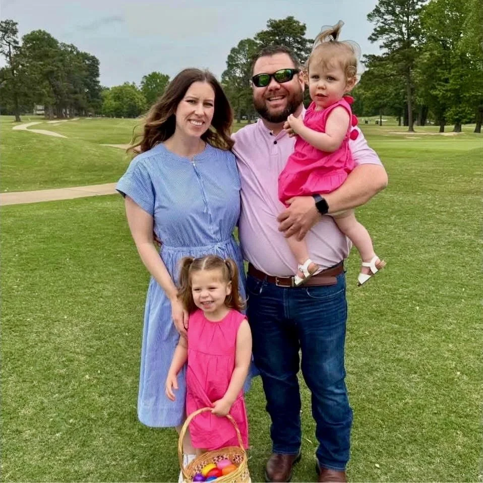 Family of four posing outdoors on a golf course, with two young girls in pink dresses, smiling parents, and a man wearing sunglasses holding a toddler.