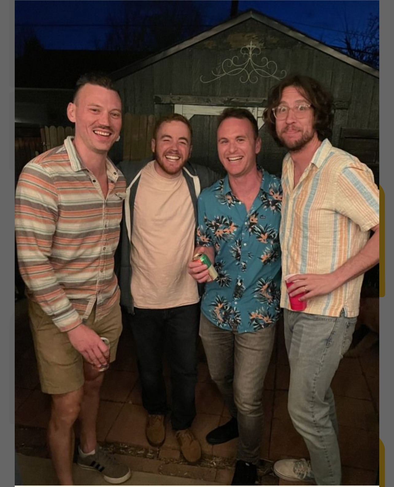 Four men standing close together at an outdoor evening gathering, smiling, with a wooden building in the background, holding drinks.