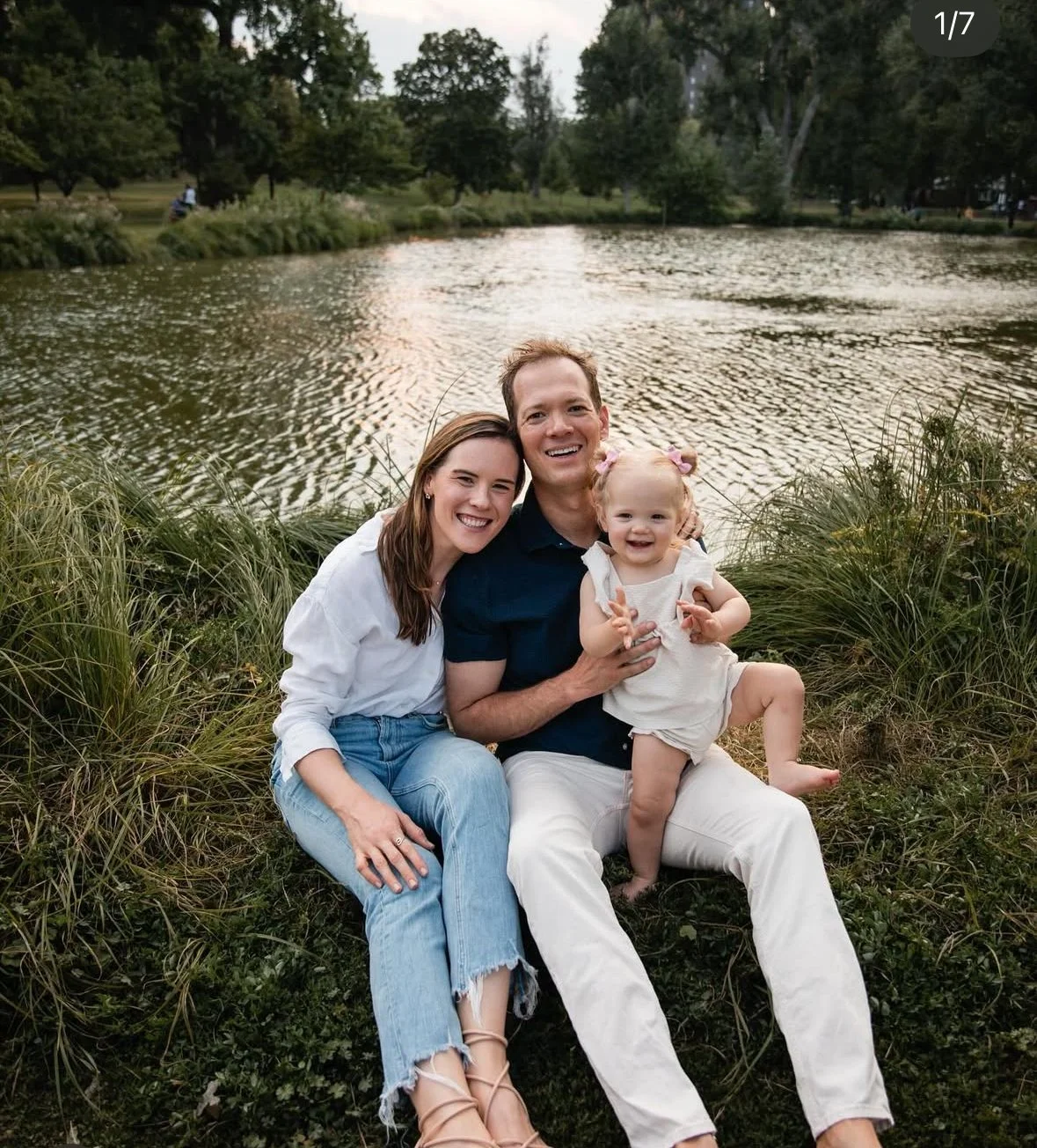A happy family of three sitting by a pond in a park, smiling at the camera. The family consists of a woman, a man, and a young girl. The woman has long brown hair, the man has short brown hair, and the girl has blond hair tied in pigtails with pink bows. The background shows trees and a peaceful outdoor setting.