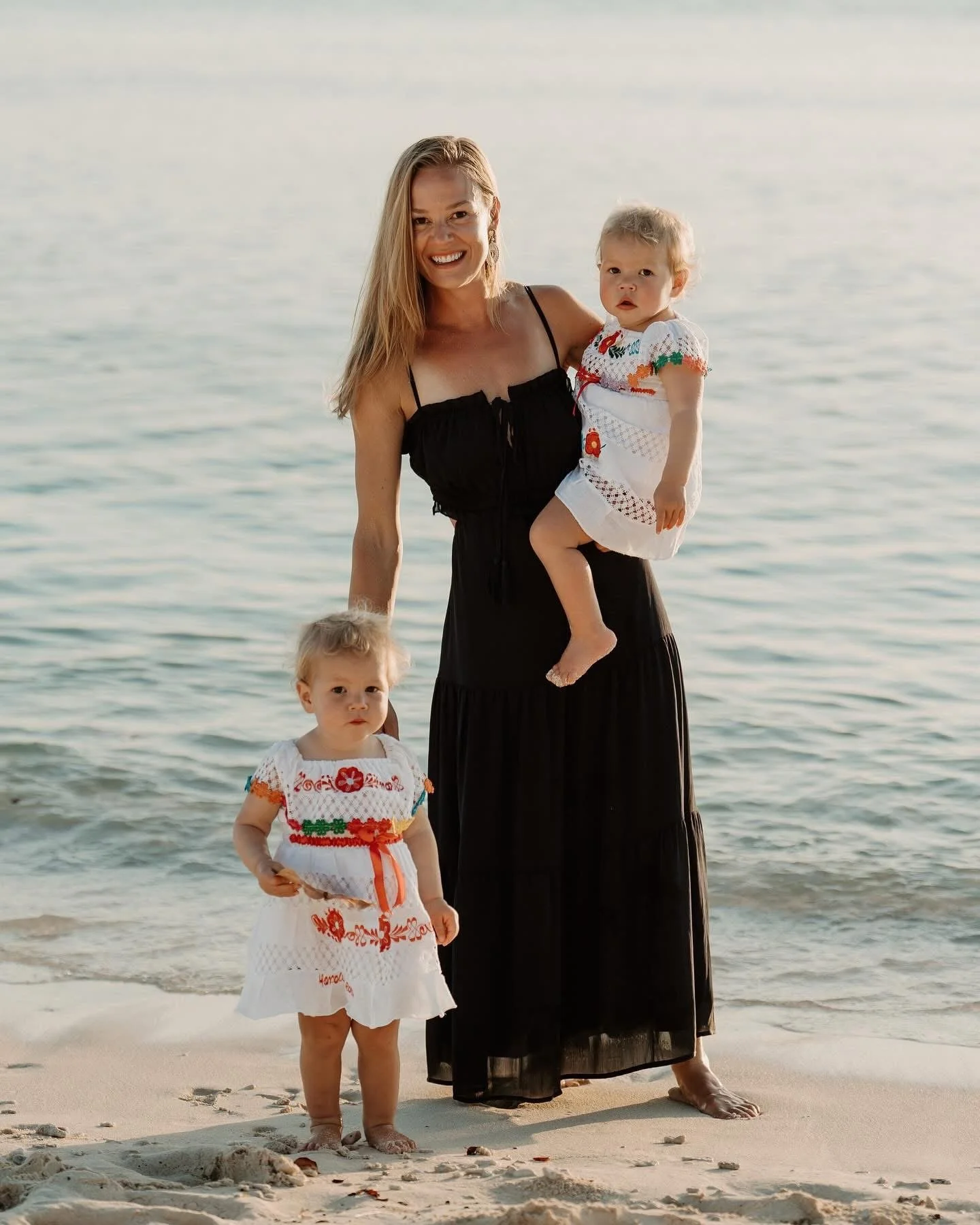 A woman in a black dress smiling on the beach, holding a young girl in a white embroidered dress, with a toddler in a similar dress standing on the sand nearby. The background shows calm water and a clear sky.