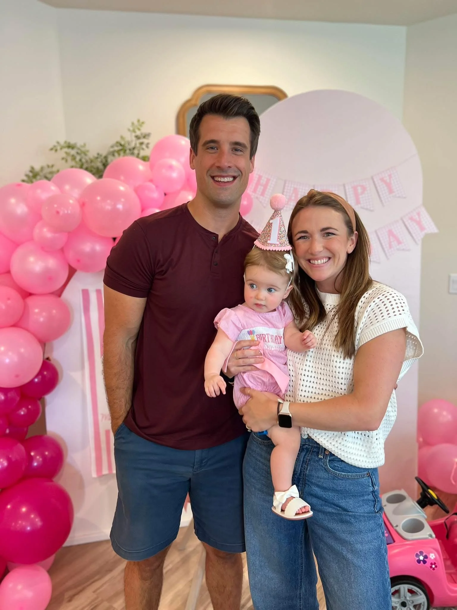 A smiling family of three, a man, a woman, and a young girl, celebrating a birthday in a decorated room with pink balloons and a 'Happy Birthday' banner. The girl is wearing a pink dress and birthday hat.