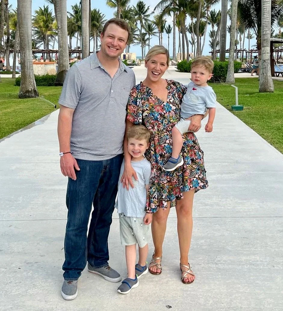 A family of four standing on a sidewalk at a tropical beach resort with palm trees in the background. The father, mother, and two young boys are smiling and dressed casually for warm weather.