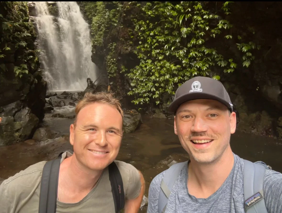 Two smiling men with backpacks taking a selfie in front of a waterfall surrounded by lush green foliage.
