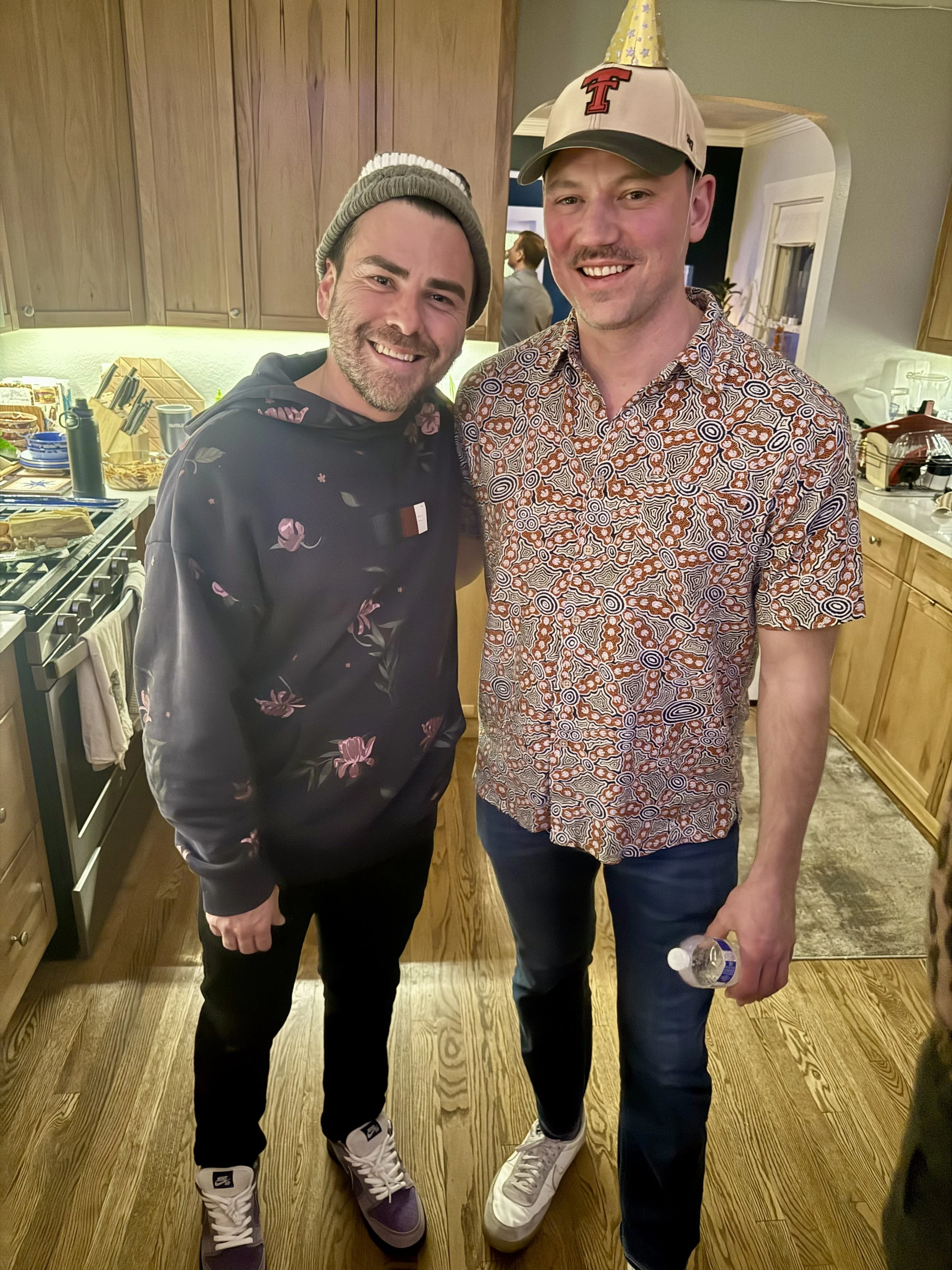 Two men smiling at a party, standing in a kitchen. One wearing a beanie and floral hoodie, the other wearing a patterned shirt and a baseball cap with an 'T' and a party hat, holding a water bottle.