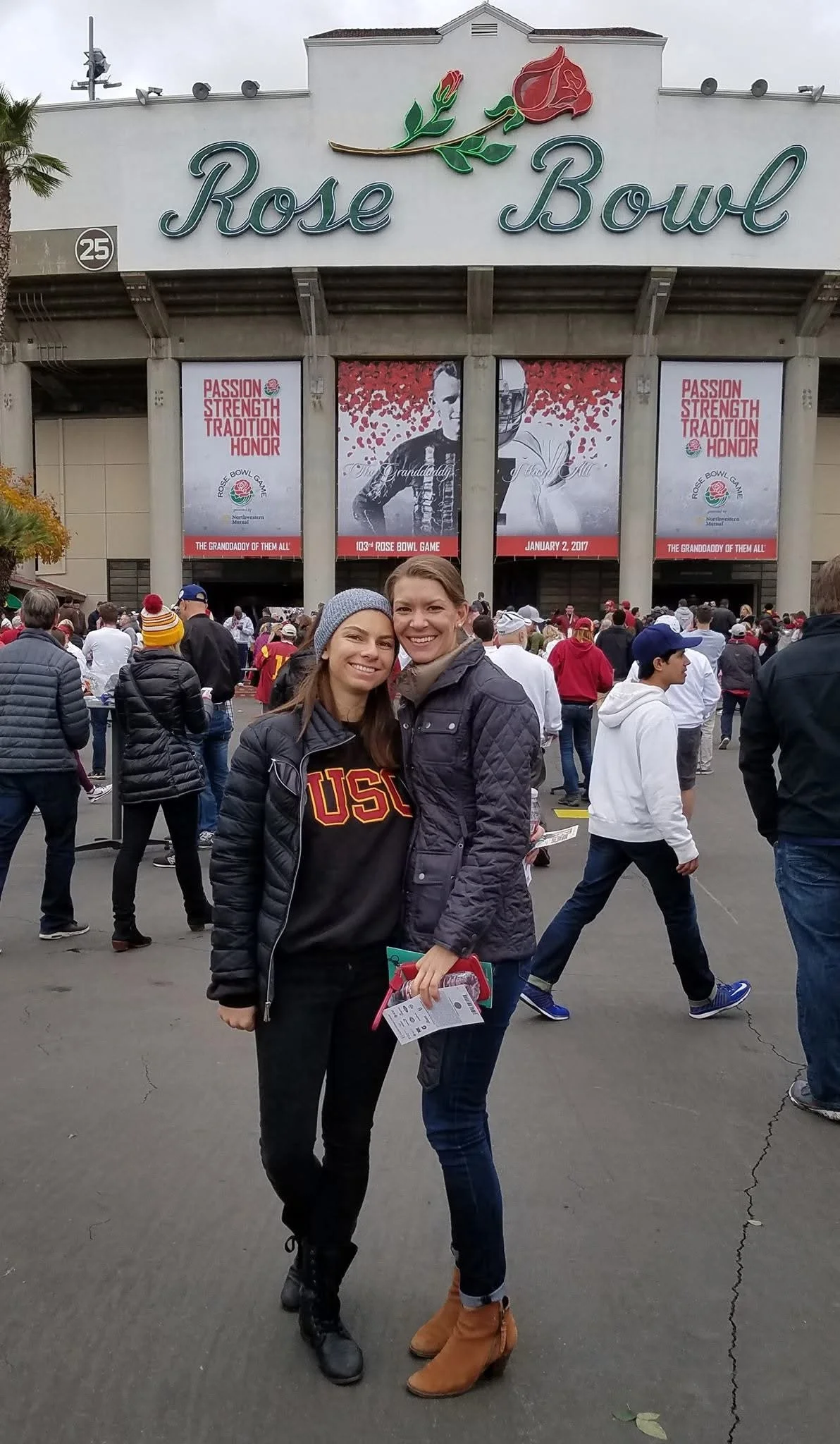 Two women smiling and standing together outside the Rose Bowl during a sports event. The background shows a crowd of people, large banners, and the Rose Bowl sign.