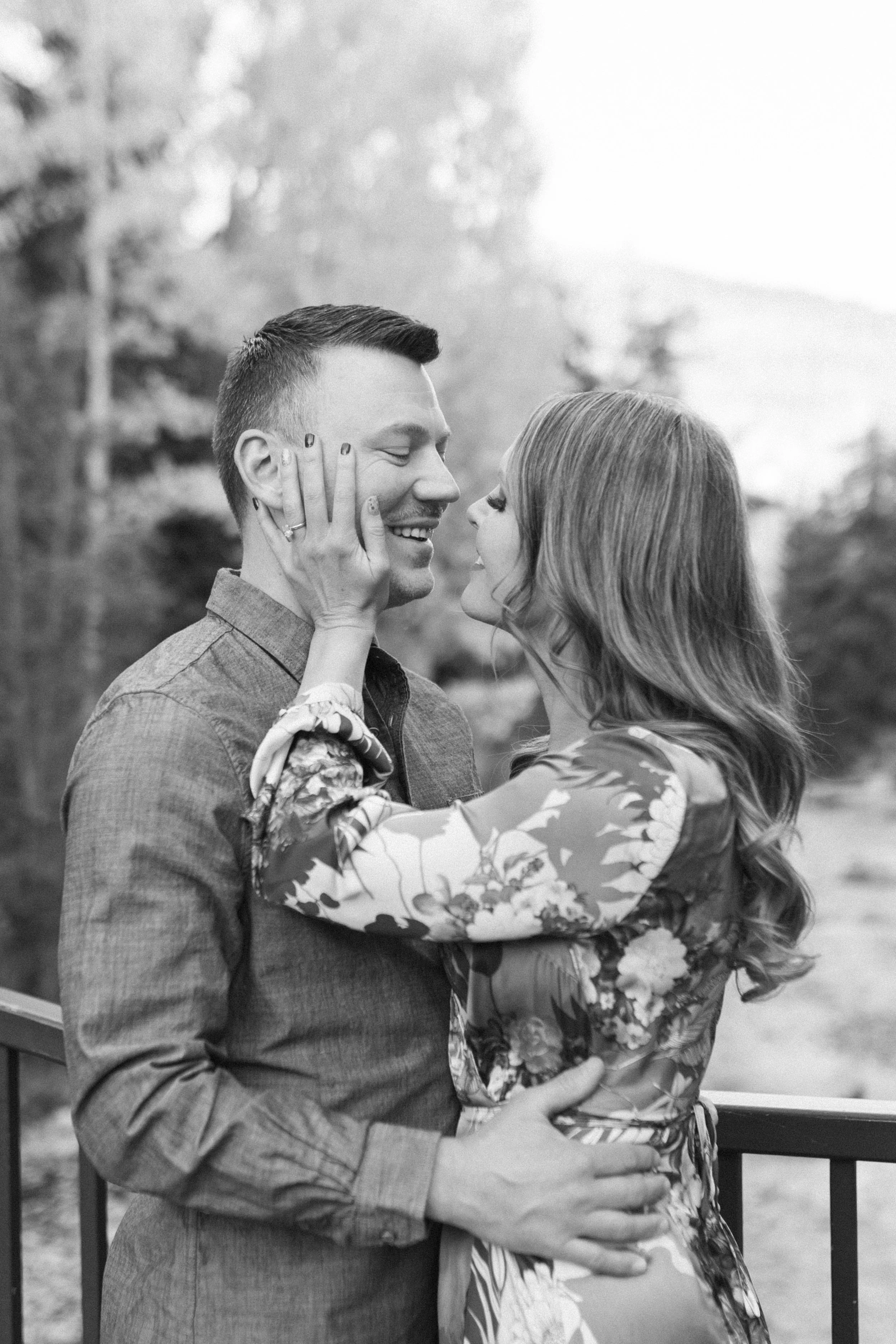 A black-and-white photo of a couple embracing outdoors, smiling at each other, with trees and a blurred landscape in the background.