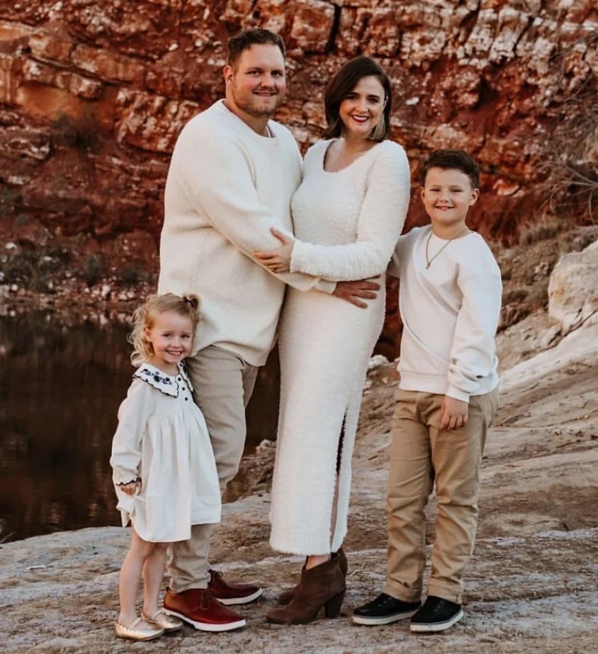 A family of five posing outdoors near a body of water with a rocky, reddish-brown backdrop. The man and woman are standing close together, with the woman wearing a long white dress and the man in a light-colored shirt and pants. The children, a girl and a boy, stand next to them, all smiling at the camera.