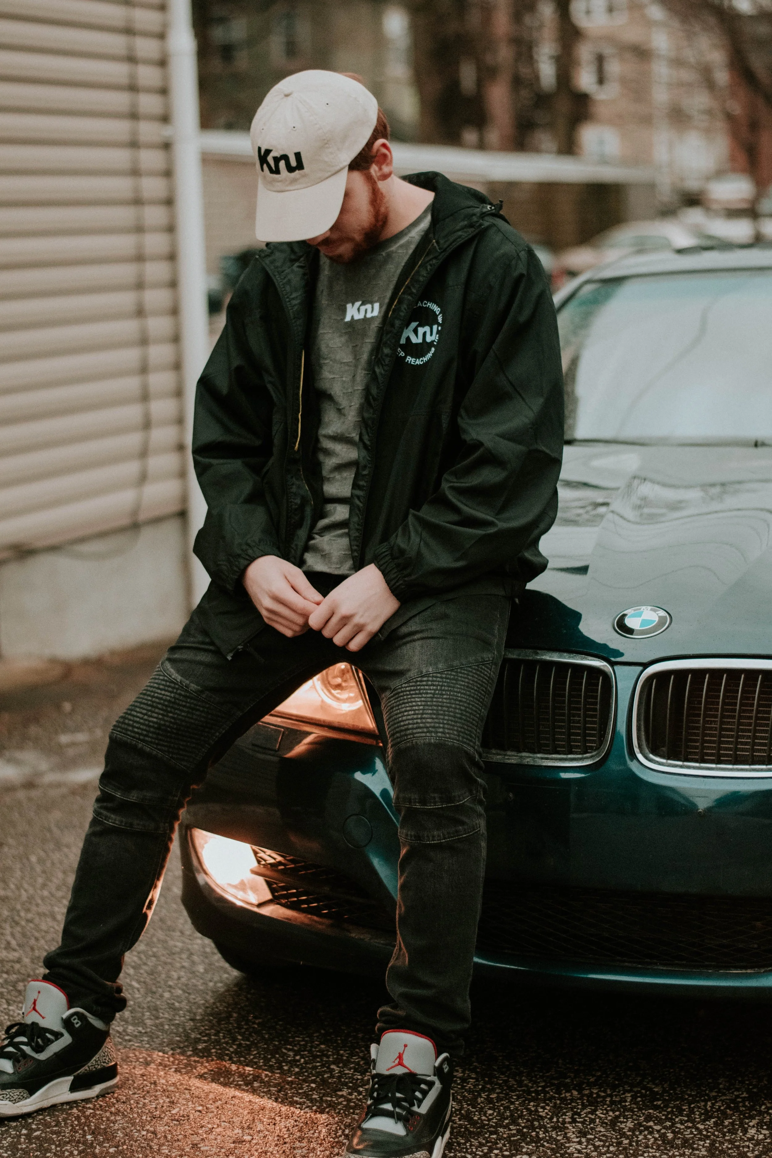 A young man with a beard sitting on the hood of a dark green BMW car parked on the street. He is wearing a white cap with black text, a black jacket, a gray T-shirt, black pants, and black and white Air Jordan sneakers. The background includes a beige building, a wooden fence, and some trees.