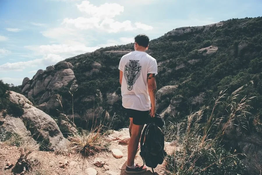 A man with dark hair, wearing a white t-shirt with a tiger graphic on the back, black shorts, and sneakers, stands on a rocky trail in a desert landscape with hills and large rocks, holding a black backpack.