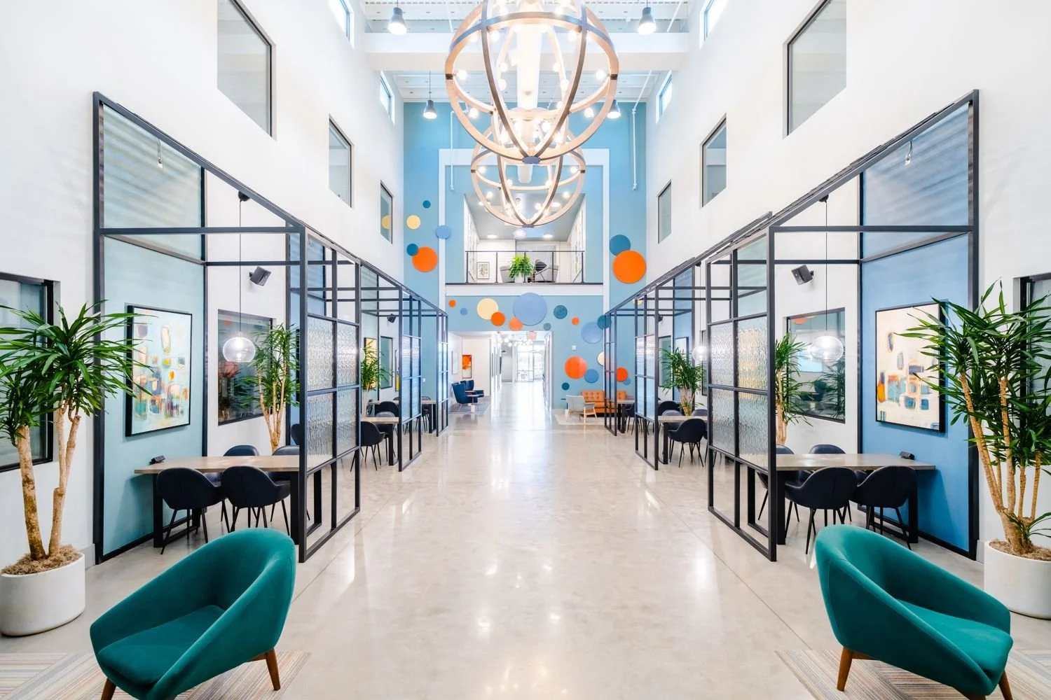 Bright, modern two-story office lobby with blue walls, colorful circular art decor, hanging wooden light fixtures, and seating areas with teal chairs and potted plants