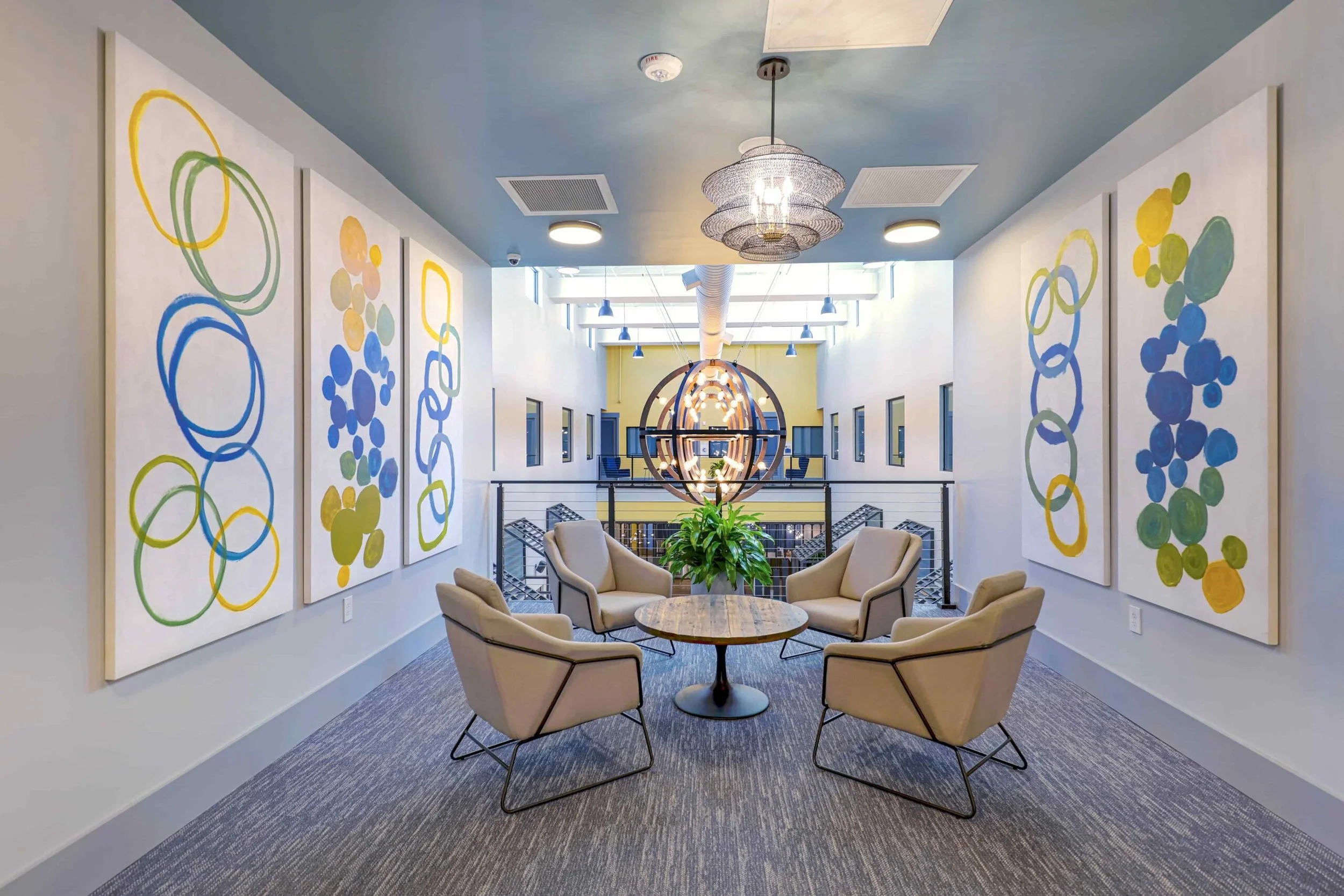 A modern interior space with six beige armchairs arranged around a round wooden table with a green plant in the center. The walls are decorated with abstract art featuring colorful circles. A large pendant light hangs above, and there is an open view of a hall with blue chairs and a circular artwork hanging in the background.