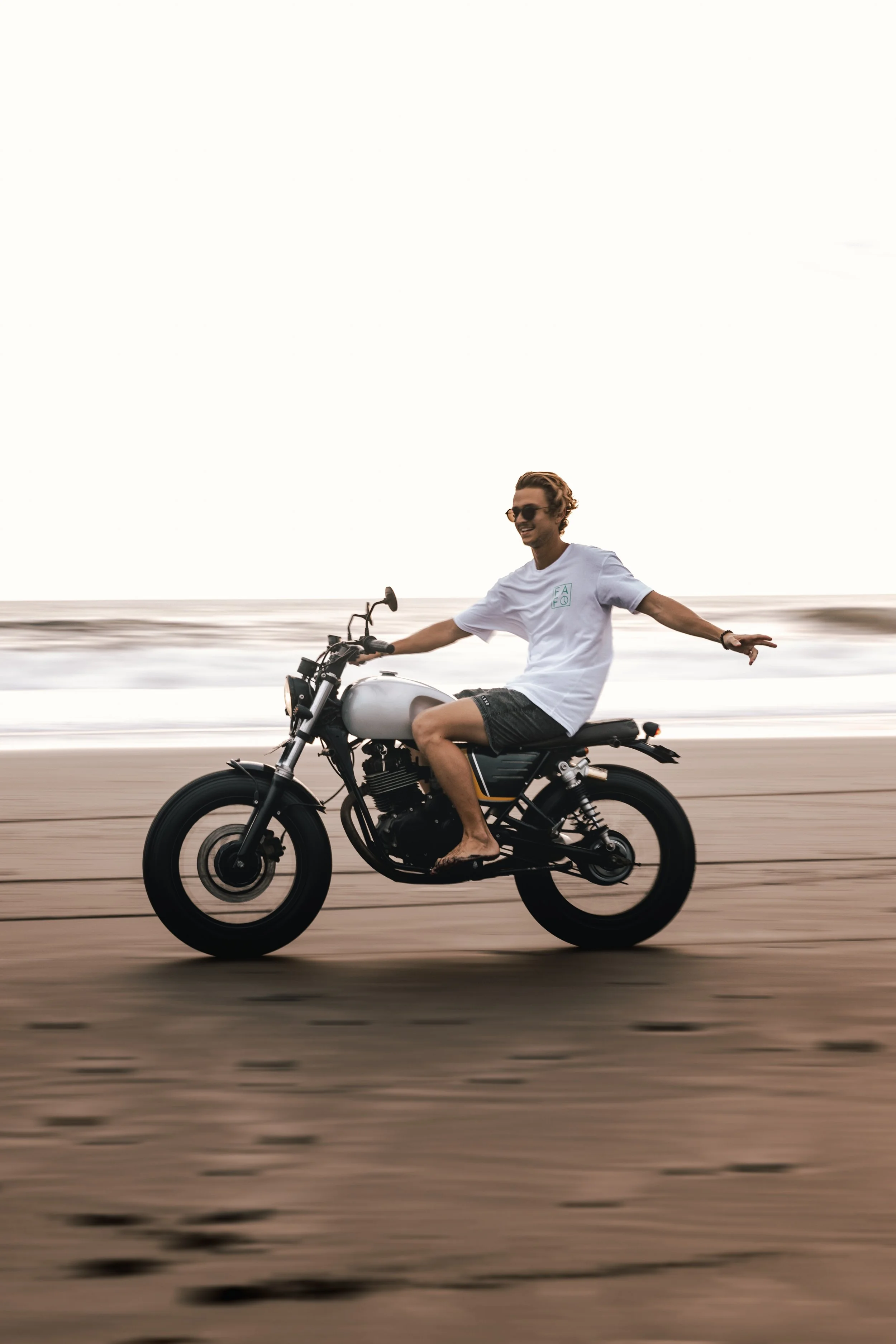 A man riding a motorcycle along the beach at sunset with his arms outstretched, wearing a white t-shirt, shorts, and sunglasses.