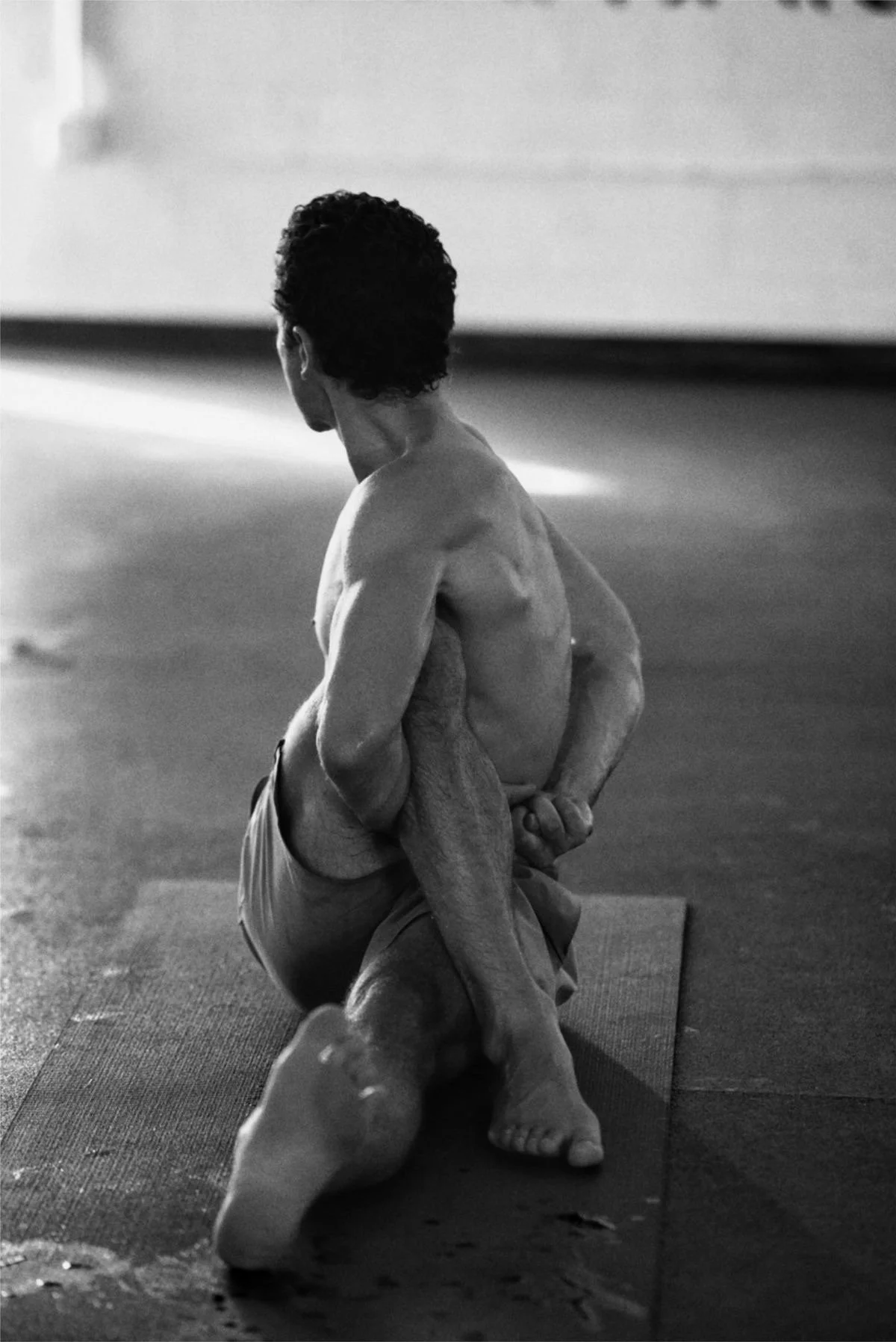 A person practicing yoga indoors, performing a kneeling spinal twist on a mat, with their back to the camera in black and white.