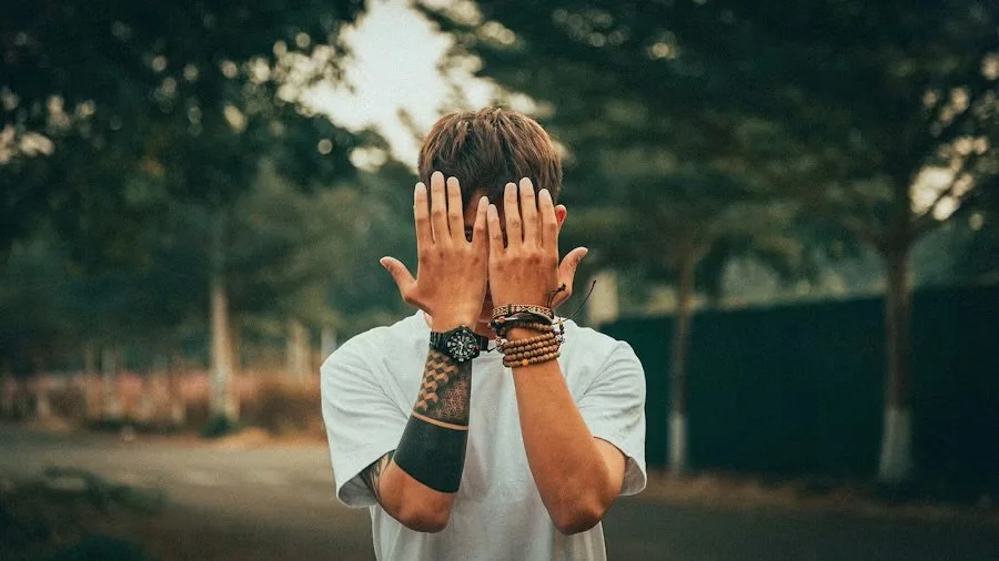 A young man with multiple bracelets and tattoos on his arms covers his face with his hands outdoors in a park-like setting.