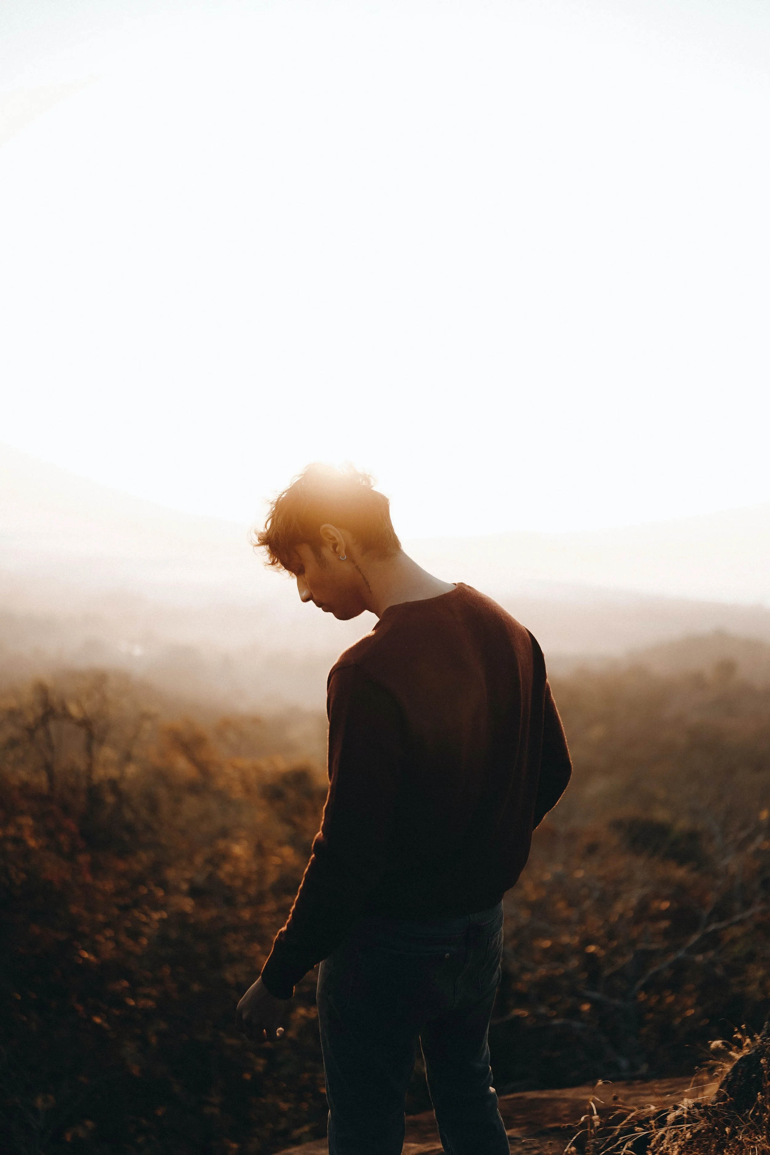 A young man stands outdoors during sunset, with a scenic landscape of trees and hills in the background.