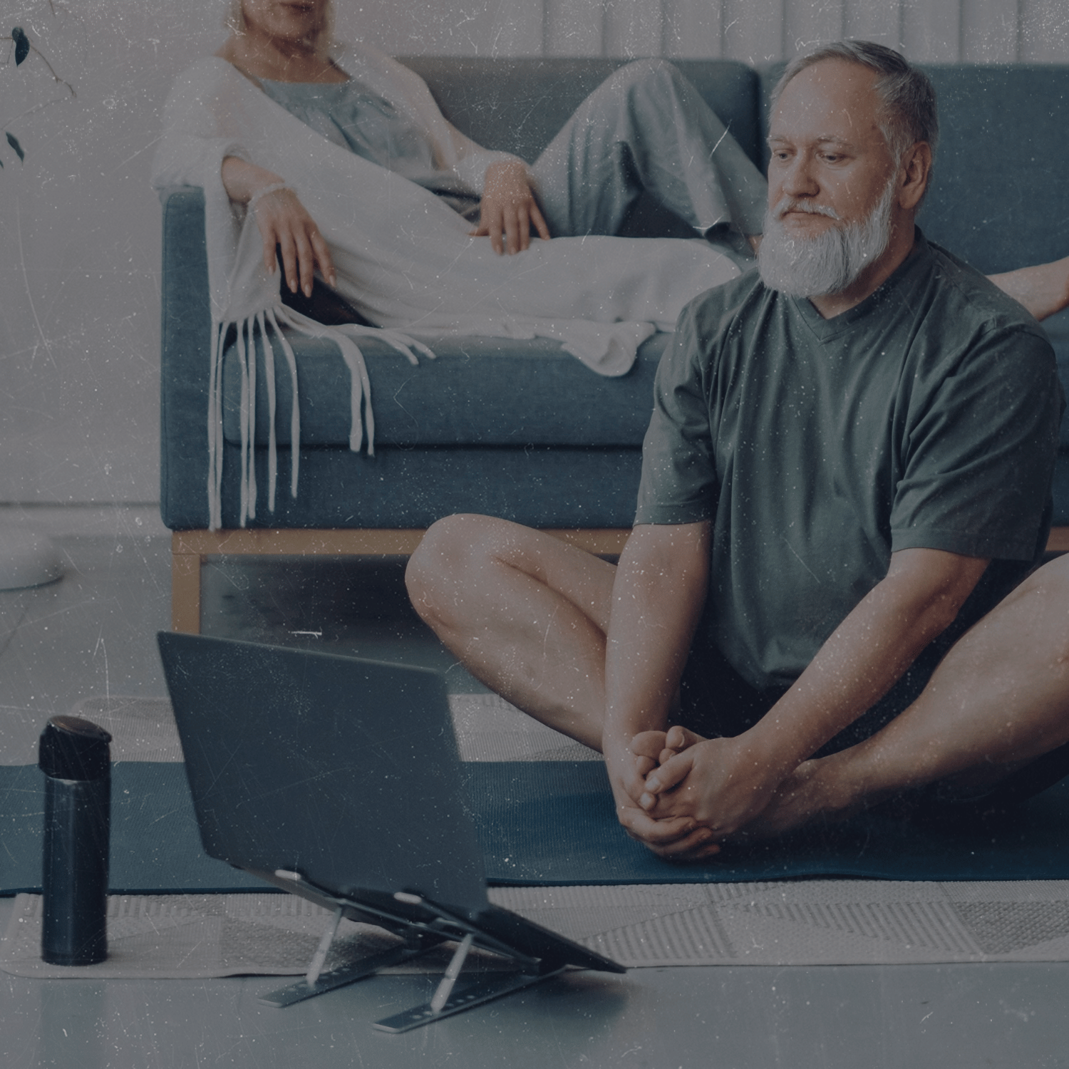 A man practicing yoga at home, sitting cross-legged on a yoga mat in front of a laptop. In the background, a woman is resting on a couch, covered with a blanket.