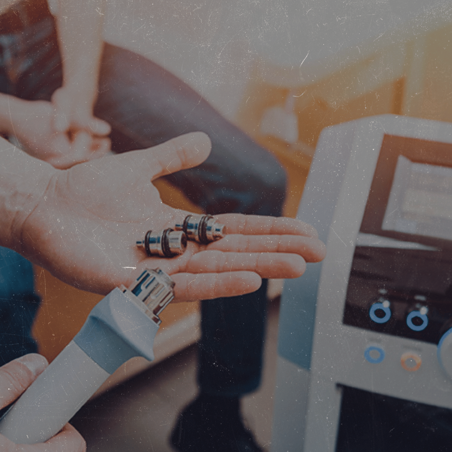 Close-up of a person's hand holding four small electronic components, with a medical device or monitor in the background.