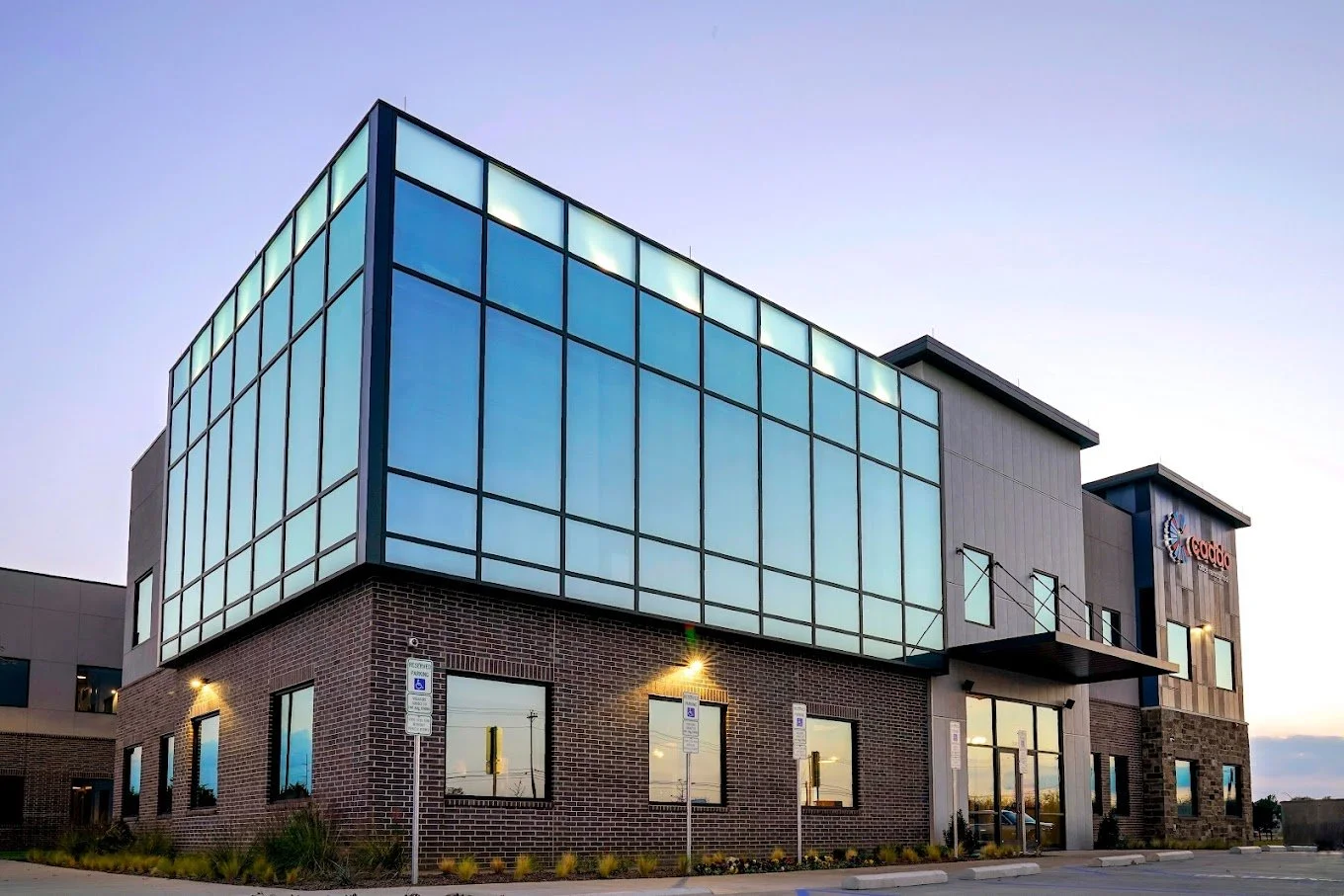 Modern commercial building with large glass windows on the upper floor, brick and gray panel facade on the lower floor, facing a parking lot with handicap parking signs, situated at dusk.