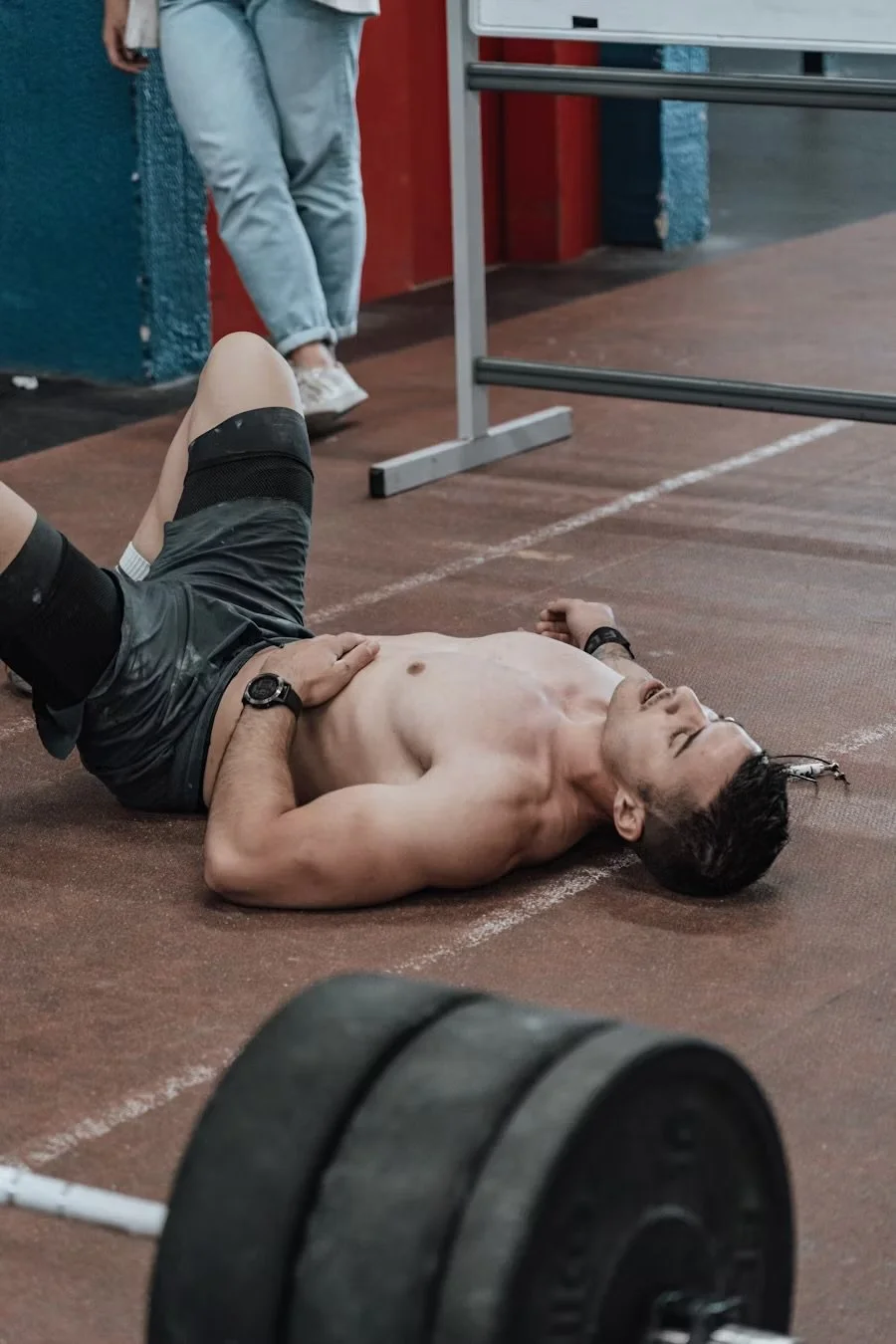 A man lying shirtless on the gym floor with a pained expression, with his hand on his stomach and a weightlifting barbell nearby.