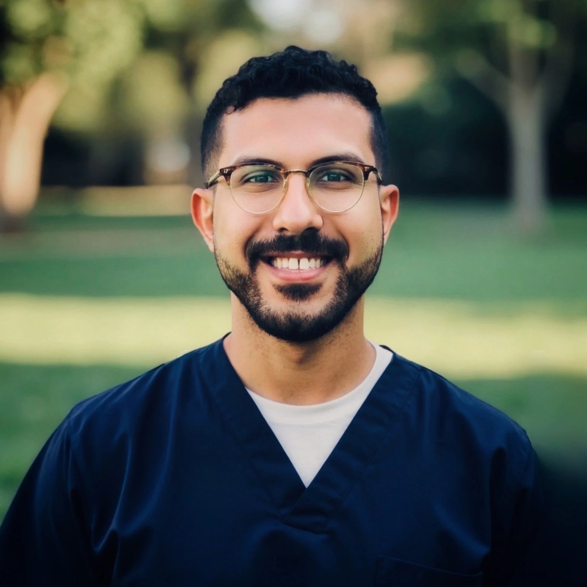 Smiling man wearing glasses and a dark medical scrubs outdoors in a park.