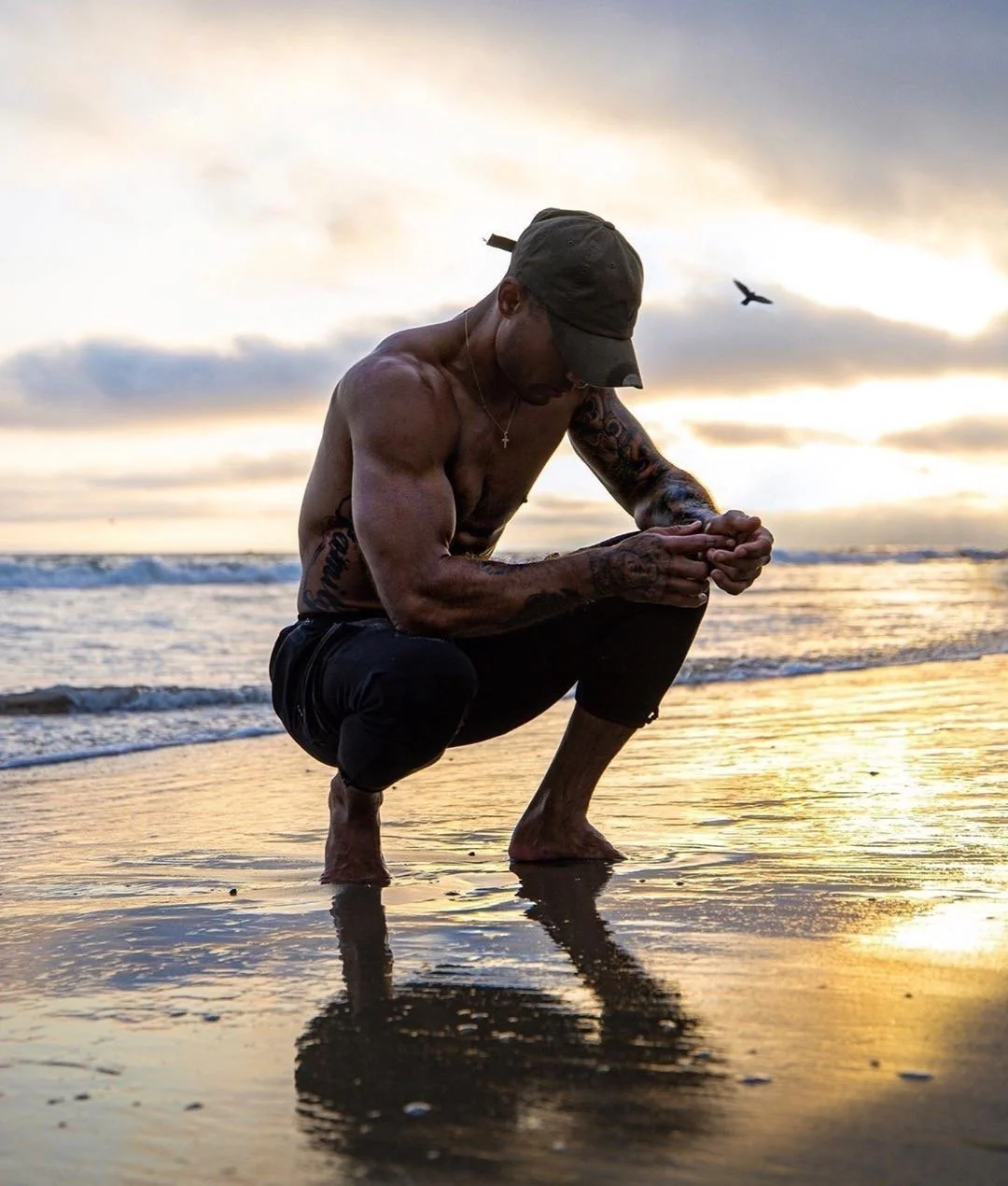 A shirtless man wearing a cap and black pants kneels on the wet sand at the beach during sunset, with his head bowed and hands clasped, reflecting his image on the water.