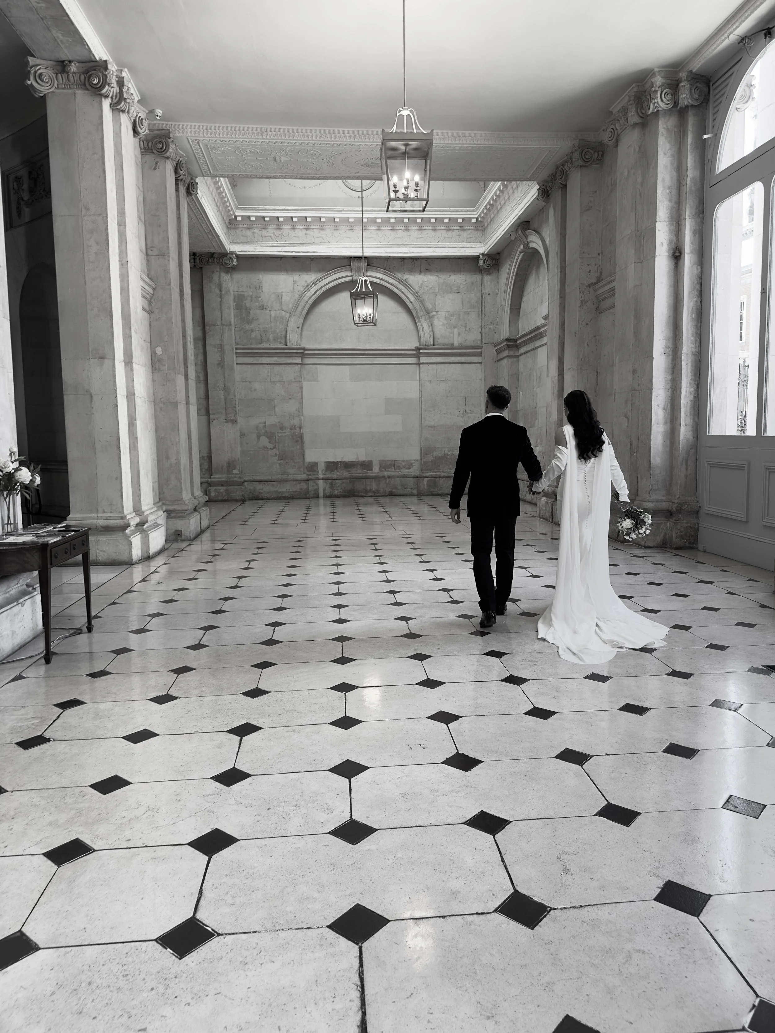 A bride and groom walking hand in hand through an elegant, historic hall with marble floors, tall windows, and ornate architectural details.
