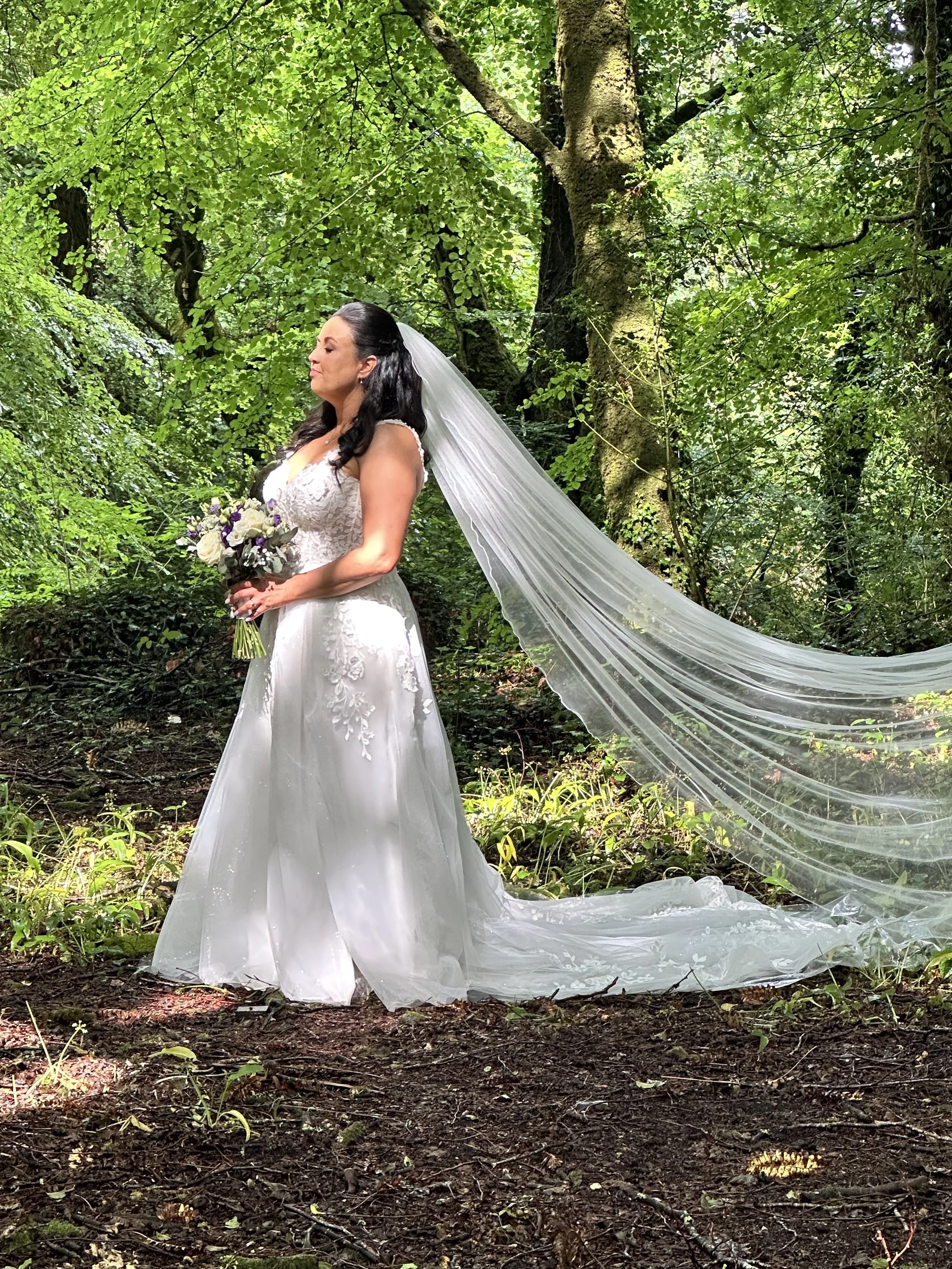 A bride in a white wedding gown holding a bouquet, standing outdoors in a lush green forest with sunlight filtering through the leaves.