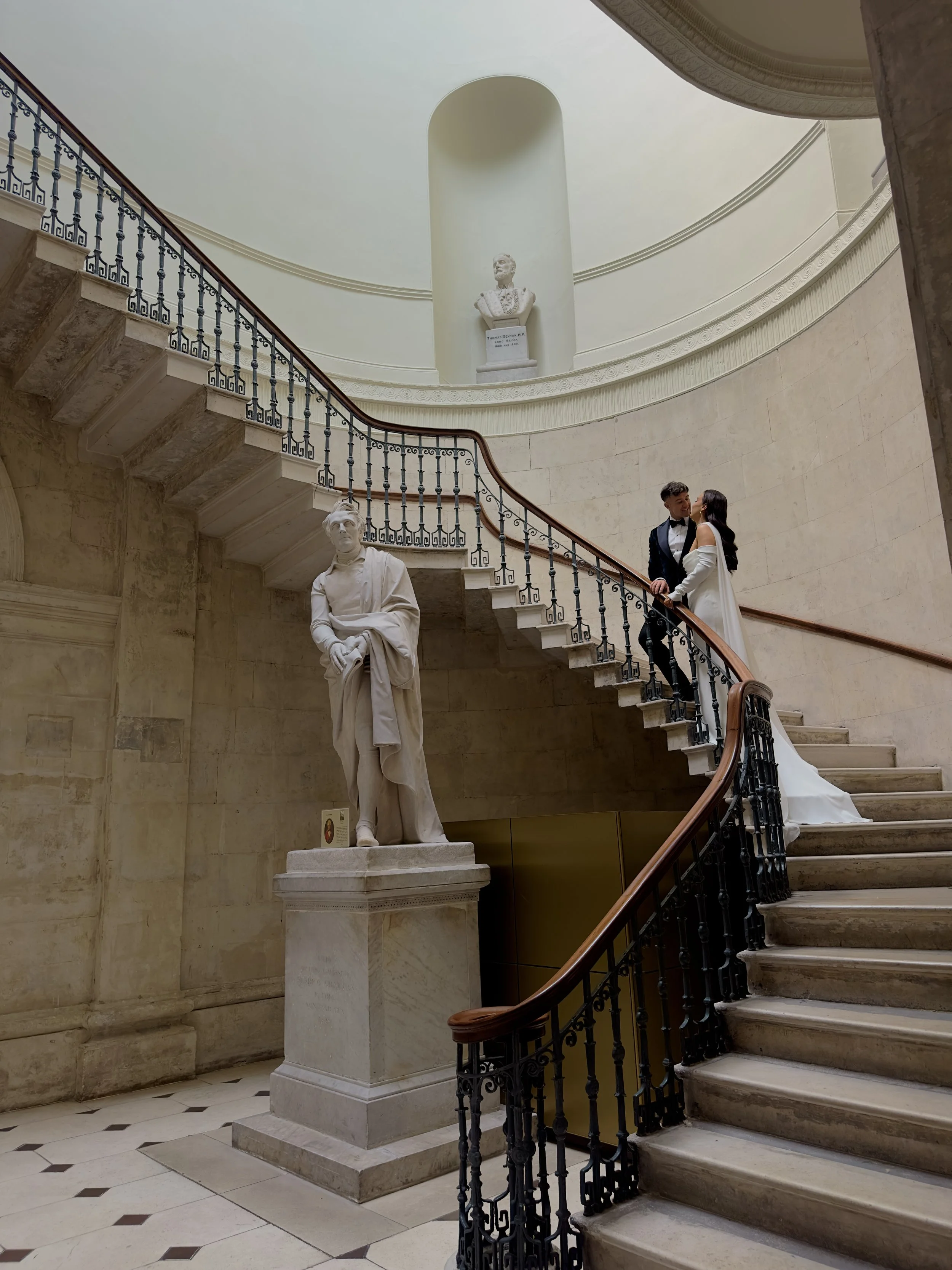 A man and woman dressed in wedding attire on a grand staircase inside a museum, with classical statues around.