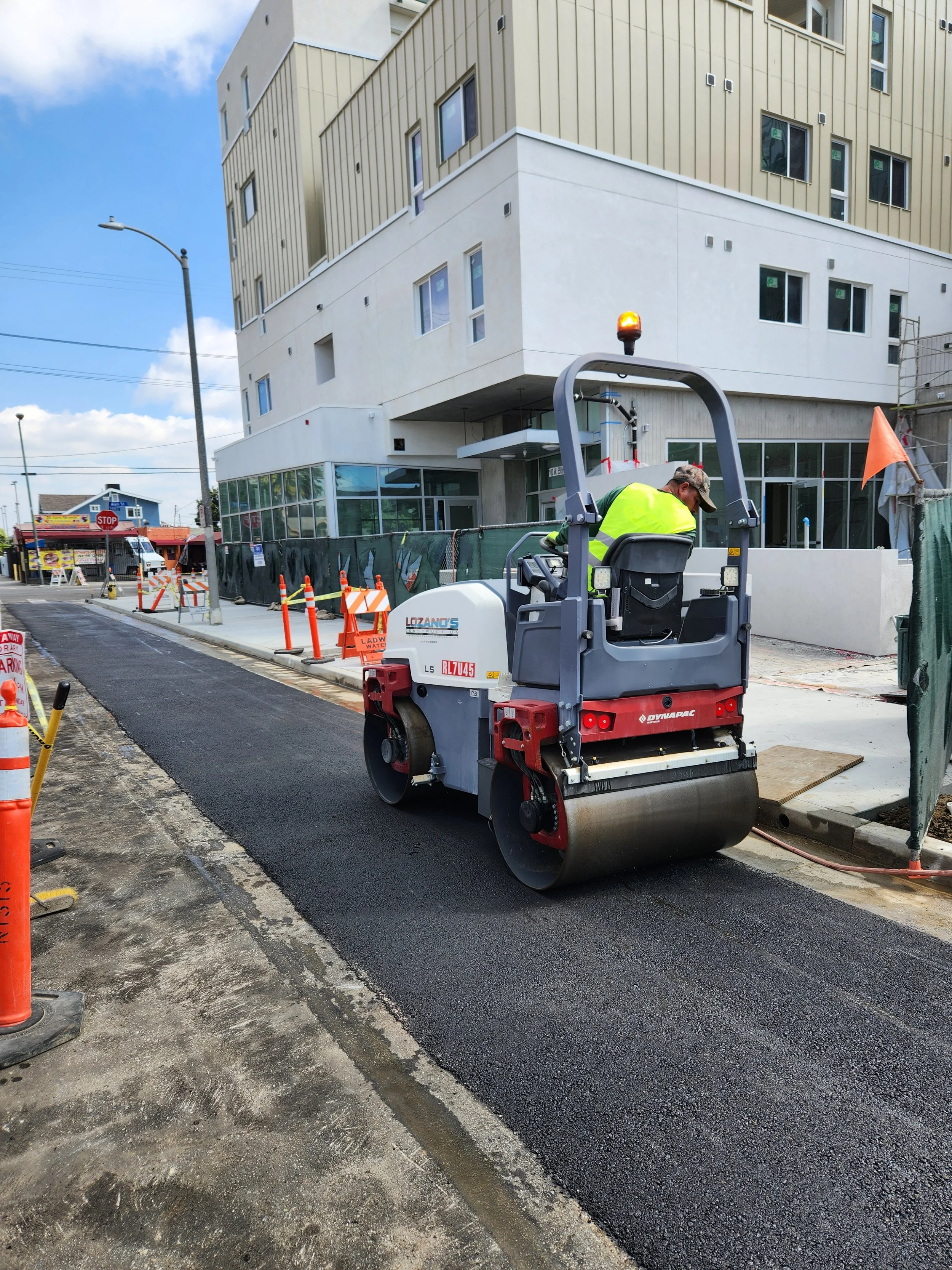 A construction worker operating a steamroller on a newly paved road in an urban area. There are construction barriers and a modern building in the background.