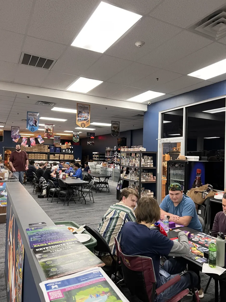 People sitting around tables playing card games at a gaming store, with shelves filled with trading card game product and posters hanging from the ceiling.