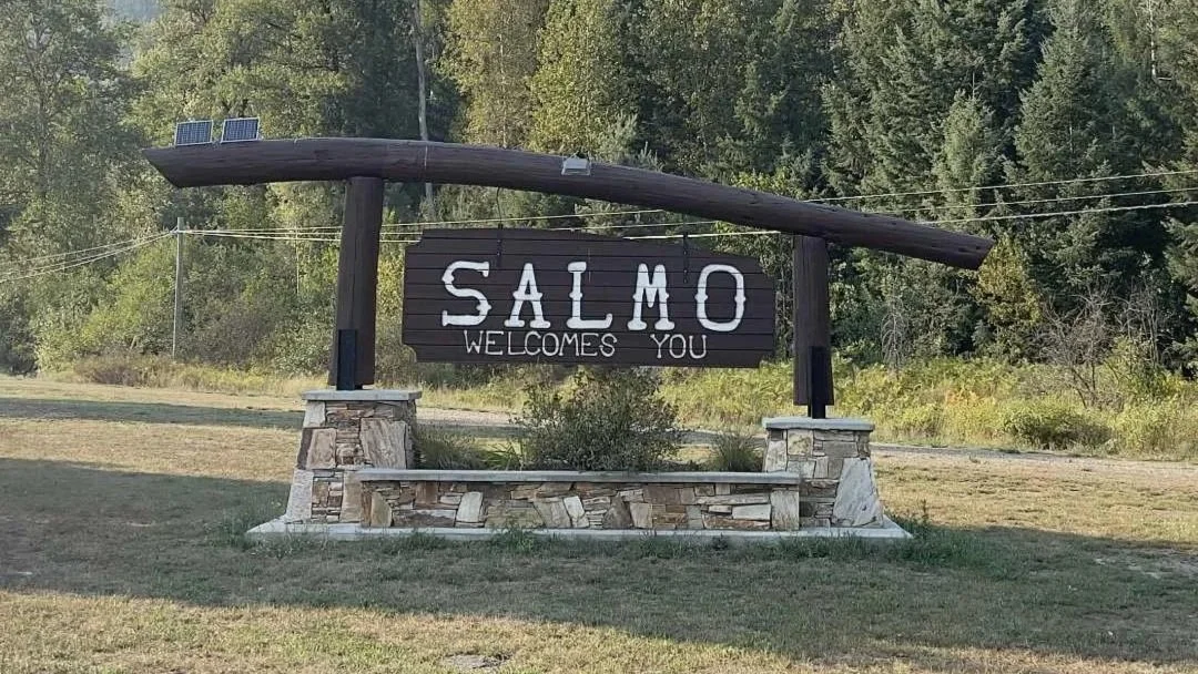 Wooden arch sits on a river stone platform. In the middle of the arch hangs a wooden sign with white lettering saying "Salmo welcomes you" The wood is dark stained.