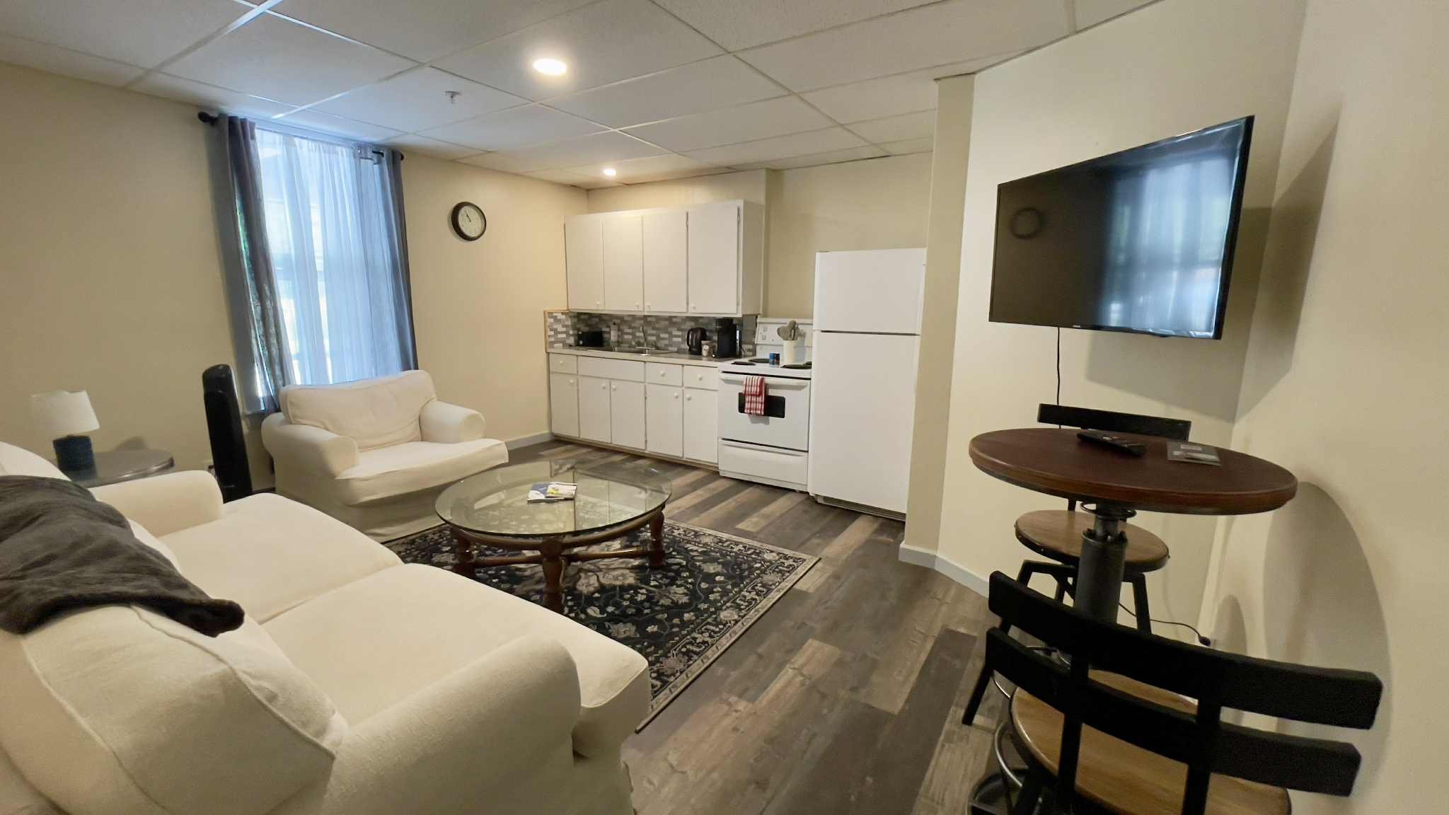 A white brocade couch on the left, glass coffee table in the centre, wooden bar stools and high table sit below the smart television on the right, and a fully stocked kitchen in the back.