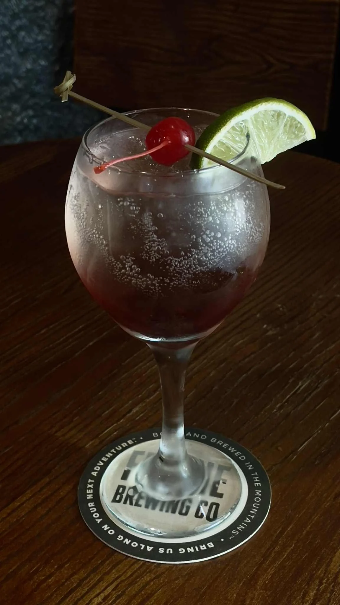A red wine spritzer cocktail with a lime and cherry garnish sits on a dark wood grain table in The Historic Salmo Hotel