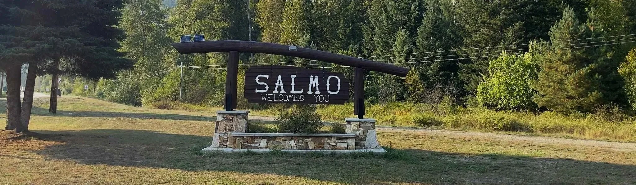 A wooden sign with white lettering saying Salmo Welcomes You at the entrance to the small mountain town in British Columbia where The Historic Salmo Hotel operates.