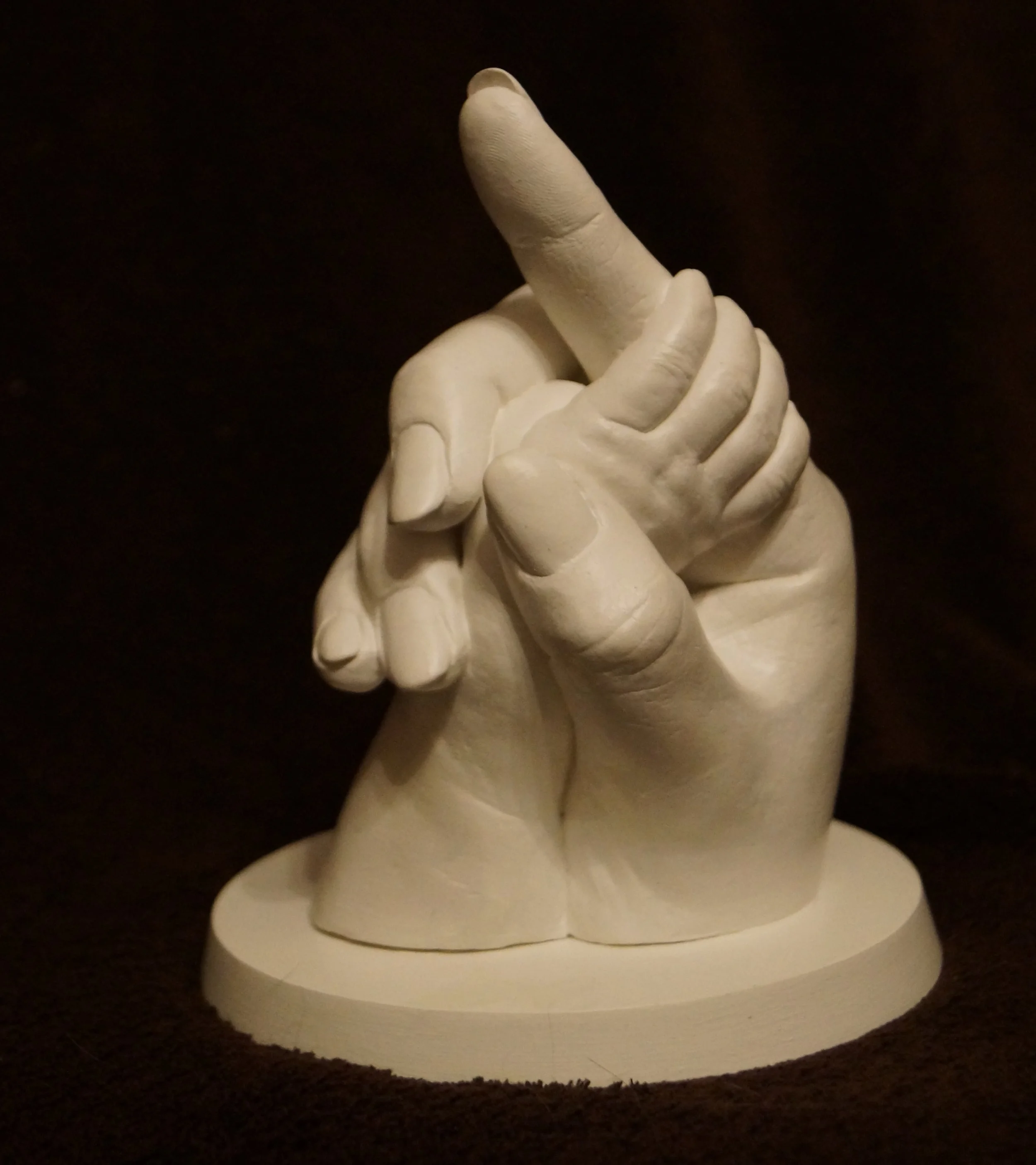 Weiße Skulptur einer Hand, die eine andere Hand hält, auf einem runden Sockel, vor einem dunklen Hintergrund.