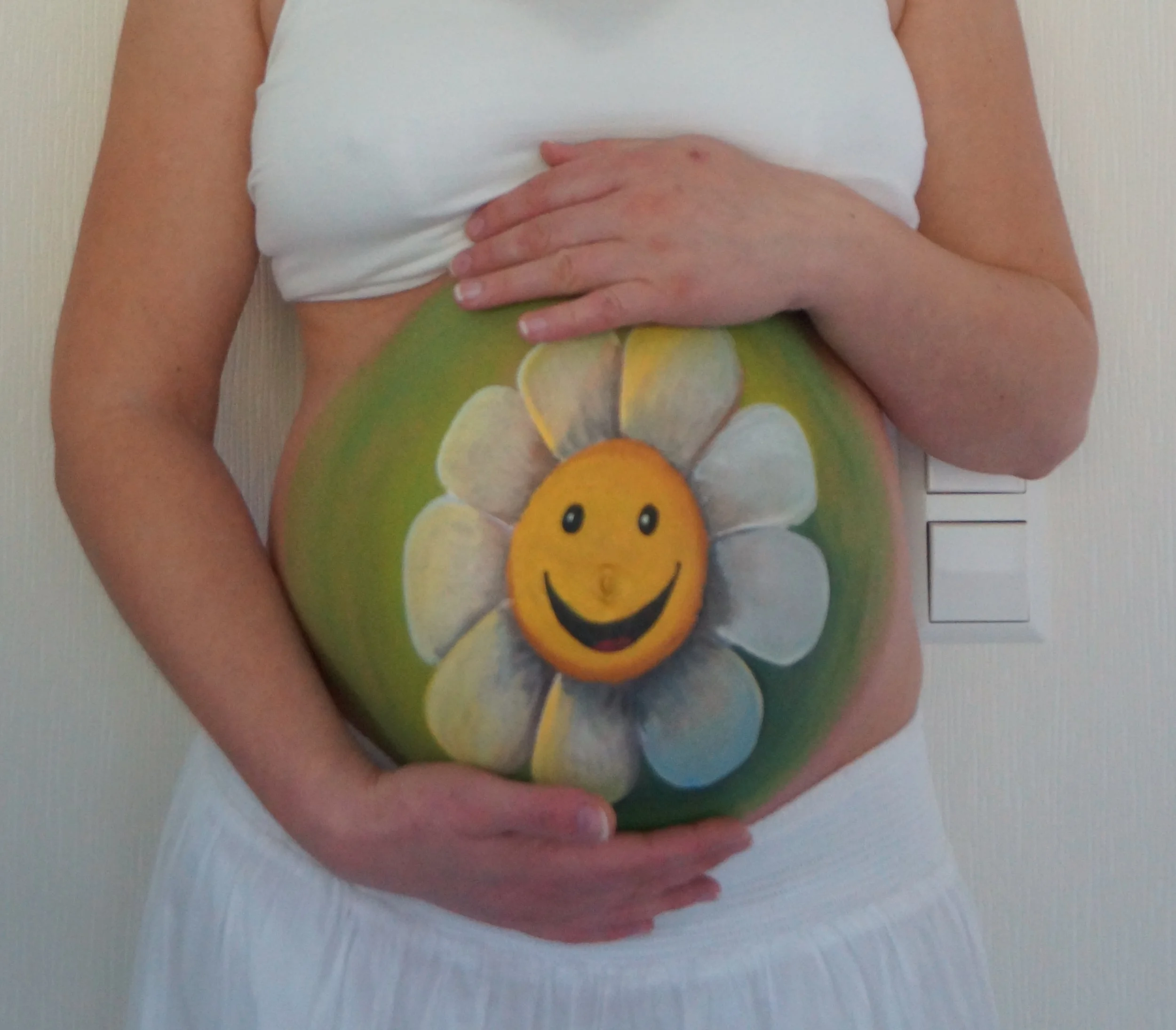 Eine Person hält die Hand auf den Bauch, der mit einem bunten Gemälde bemalt ist, das ein lachendes, orangefarbenes Gesicht inmitten von weißen Blüten zeigt.