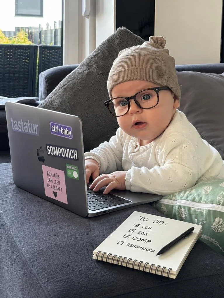 Süßes Baby mit Brille und Mütze liegt auf einem Sofa vor einem Laptop. Auf dem Laptop sind Sticker und Aufkleber, beispielsweise mit der Aufschrift "tastatur" und "ctrl+baby". Vor dem Baby liegt ein Notizbuch mit einer To-Do-Liste, die Aufgaben wie Schlaf, Essen, Spielzeug und Kuscheltiere enthält.