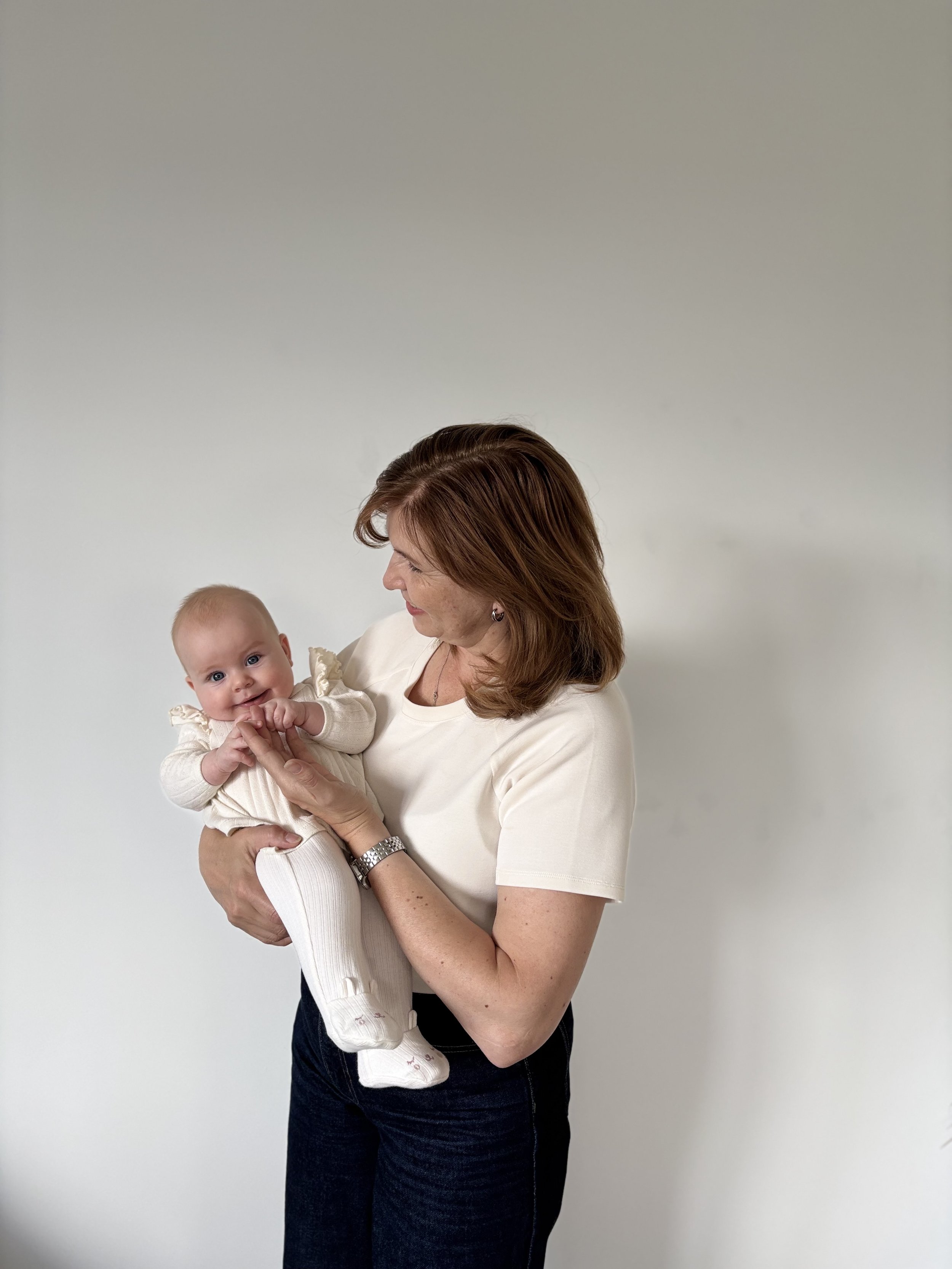 Eine Frau hält ein Baby in den Armen, beide schauen sich freundlich an. Der Hintergrund ist hell und schlicht.
