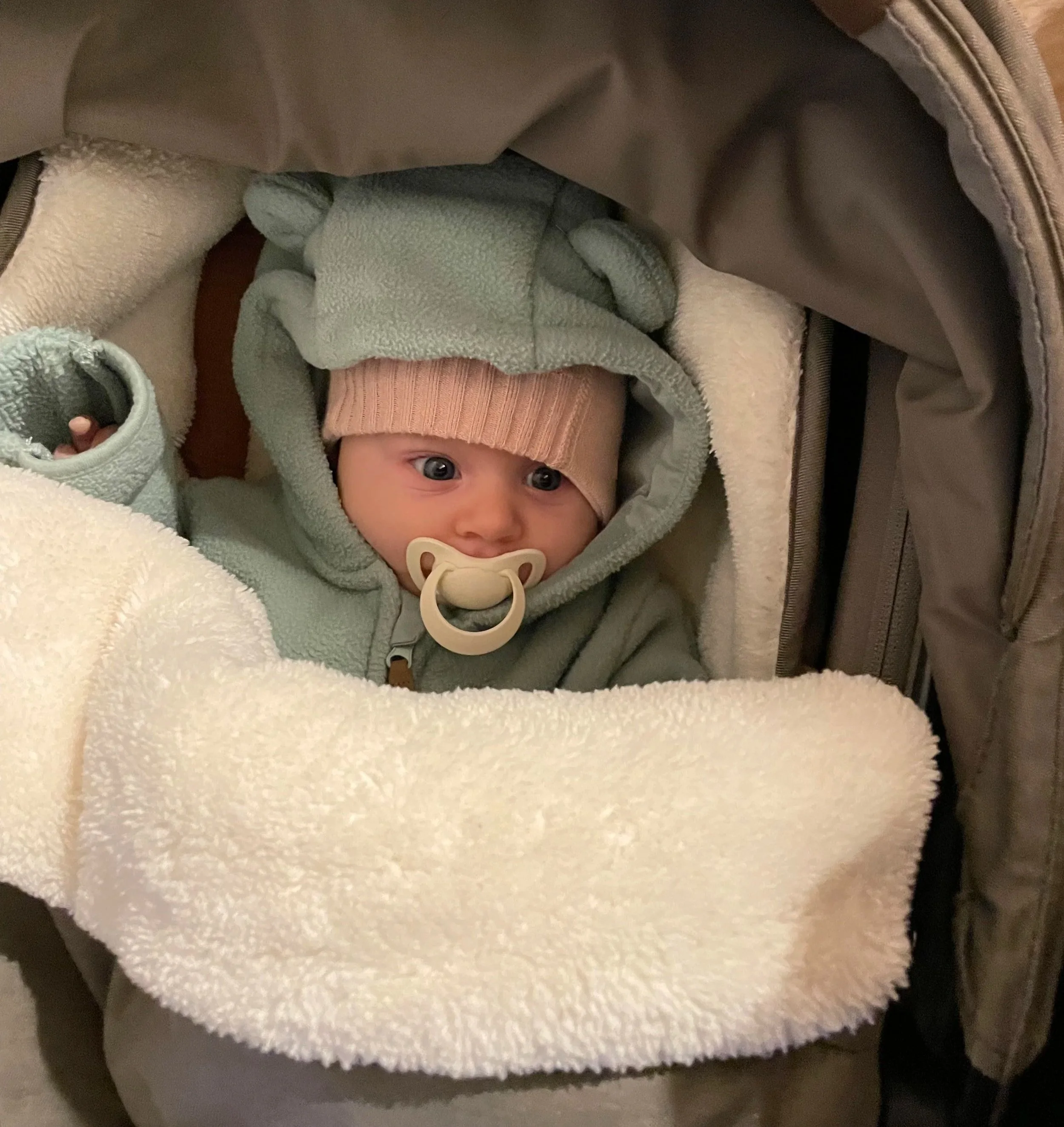 Ein Baby mit blauen Augen, eingekleidet in einen beigen Strampler und einer grünen Jacke, liegt in einem Autositz mit weißer, pelziger Decke. Das Baby hat einen Schnuller im Mund.