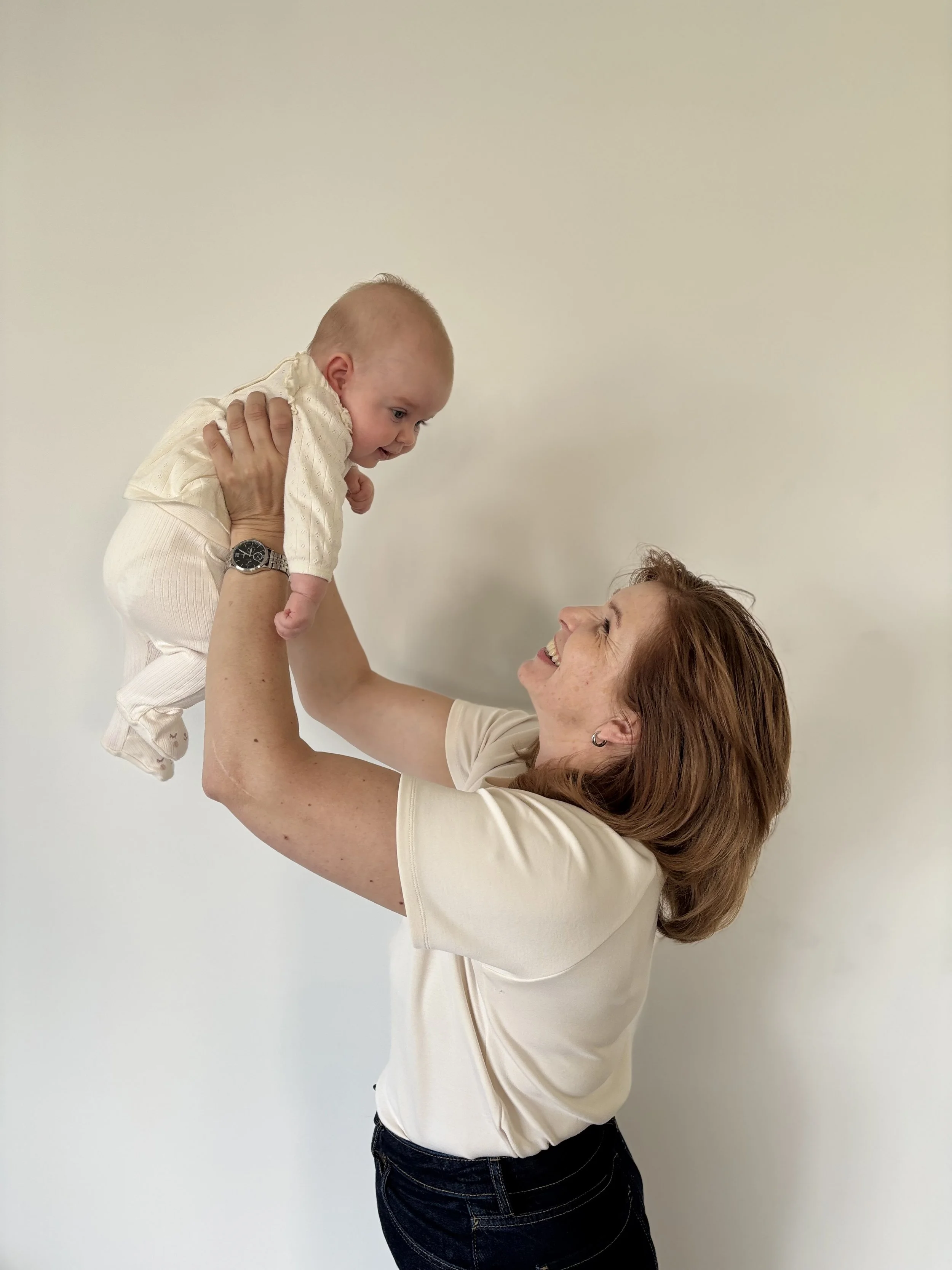 Eine Frau hebt ein Baby hoch, beide lachen und schauen sich an, vor einem weißen Hintergrund.